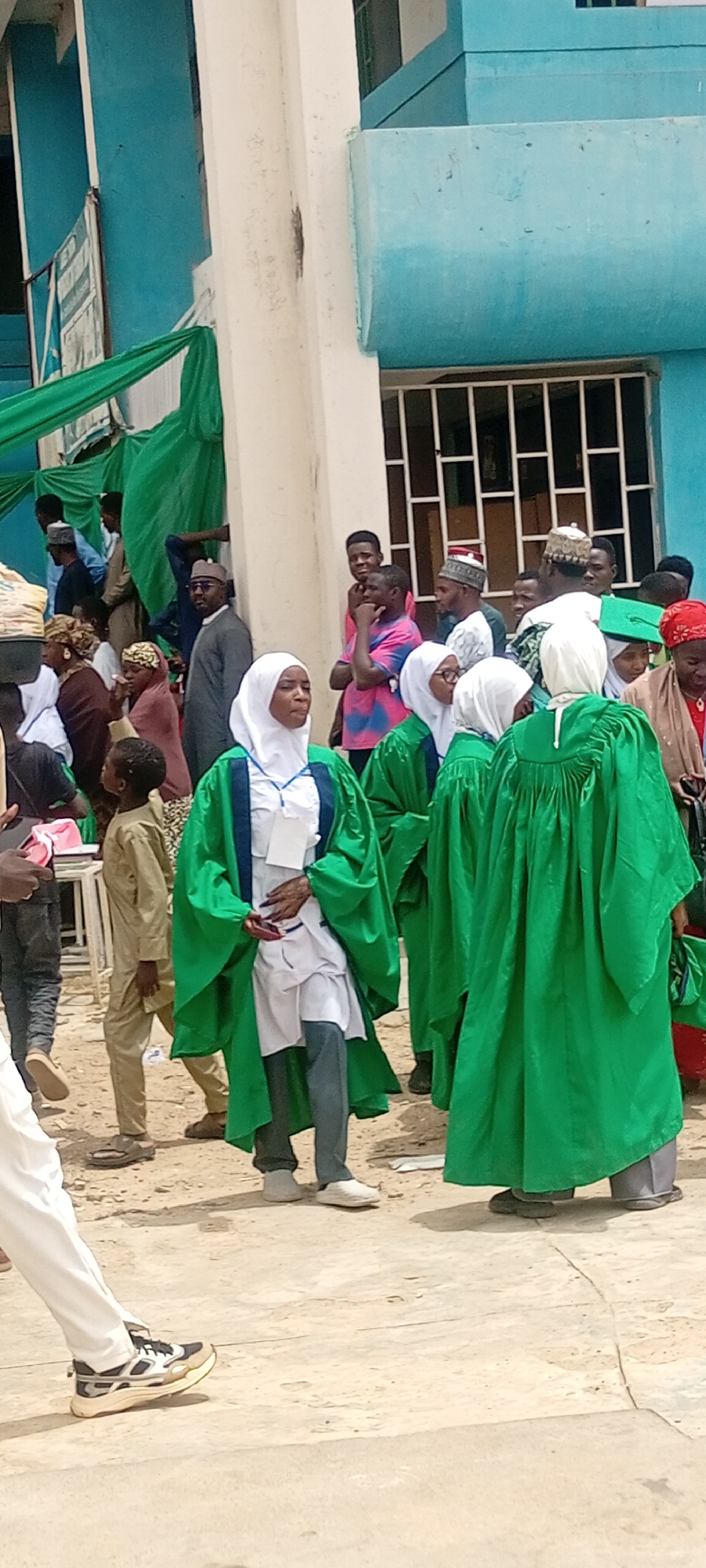 The image captures a gathering of people outside a building in Sabon Gari East, Nigeria. The building is painted turquoise and white. A group of women are wearing green gowns and white headscarves. A woman in the foreground has a white shirt, gray pants, and white shoes beneath her gown. Other people are milling around, including men in traditional attire and children. Green fabric is draped around the building. The ground is concrete.