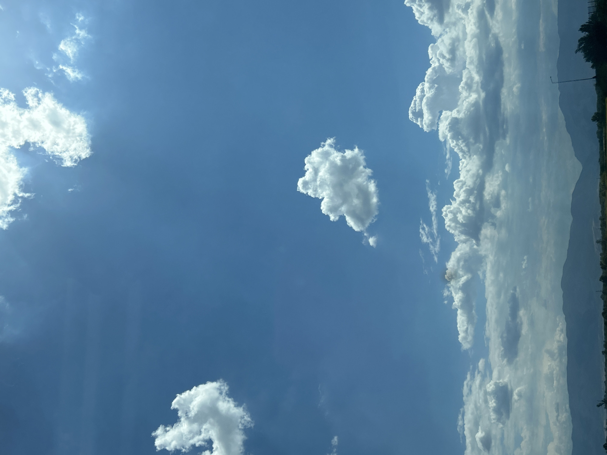 The image captures a wide-angle view of a sky filled with white, fluffy clouds against a bright blue backdrop. In the foreground, a range of dark, silhouetted mountains stretches across the horizon. Below the mountains, a strip of land is visible, with sparse vegetation and a single lamppost on the right side. The overall scene suggests an outdoor environment in a rural or semi-rural area, possibly in or around Shtarkovo, Bulgaria, during daylight hours. There are no people or specific structures that would indicate an urban setting.