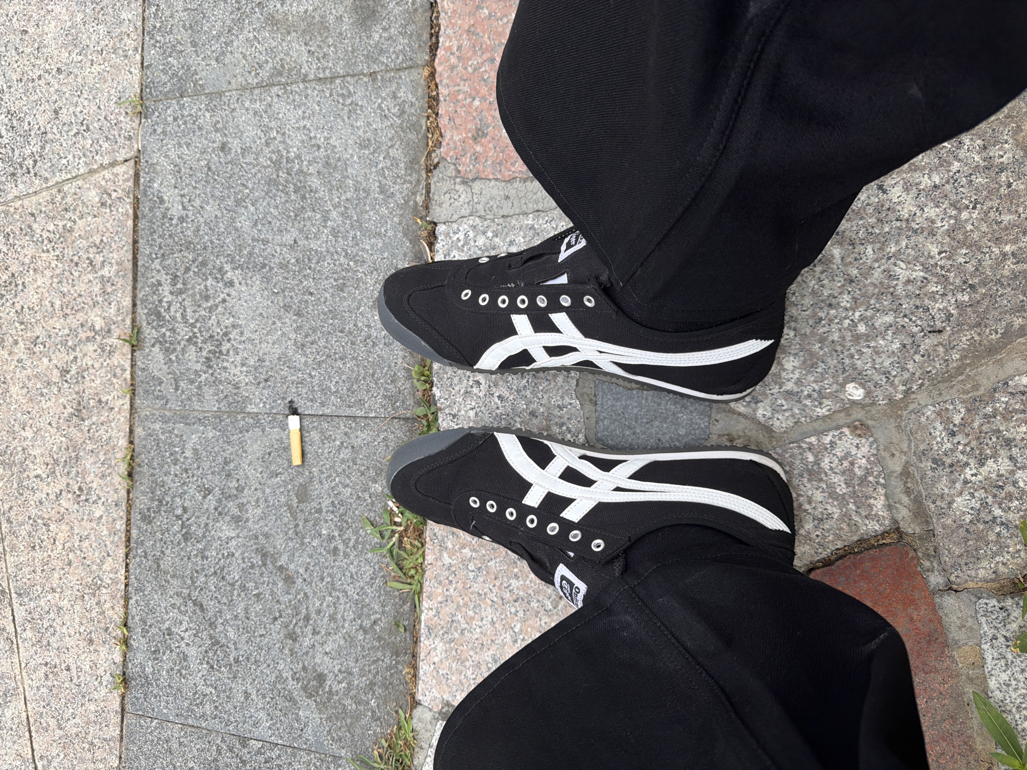 The image shows a pair of black and white Onitsuka Tiger sneakers on a person's feet, seated on a stone-tiled outdoor surface. A cigarette butt is visible on the ground between the two shoes. The person is wearing black pants. The environment appears to be a public walkway, possibly in a park or street. The tiles are a mix of grey and reddish-brown granite.