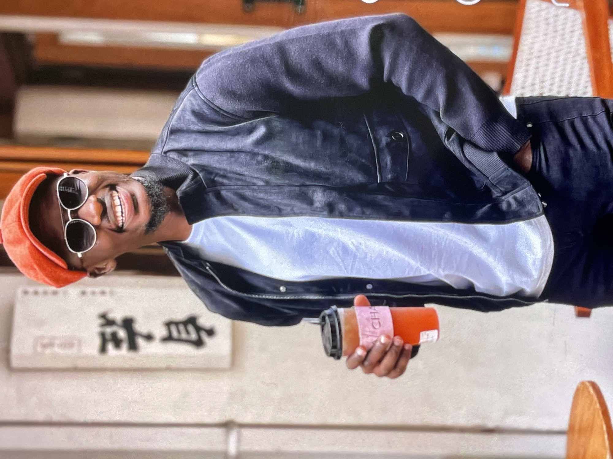 A Black man with a beard and sunglasses, wearing an orange cap, a black jacket over a white t-shirt, and black pants, stands outdoors holding a beverage in a plastic cup. He is smiling and looking to his right. In the background, a sign with Asian characters is visible on a light-colored wall. The man's left hand is in his pocket, and his right hand holds the drink. The scene appears to be in an urban setting, possibly near a cafe.