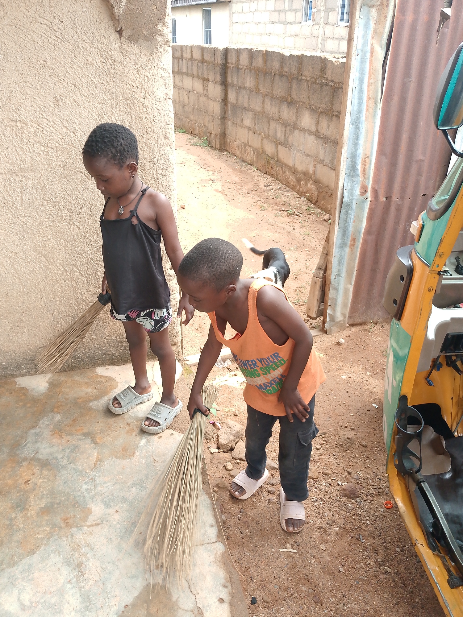 Two children, a girl and a boy, are standing and sweeping. The girl wears a black tank top and patterned shorts, and the boy wears an orange tank top and dark pants. They are both wearing sandals. Each is holding a broom made of bundled plant fibers. A black and white dog stands behind the boy. A three-wheeled vehicle, colored yellow and green, is parked to the right. The ground is a mix of concrete and dirt. Walls of concrete blocks and corrugated metal form the backdrop. The scene appears to be outdoors in a residential area in Jos, Nigeria.