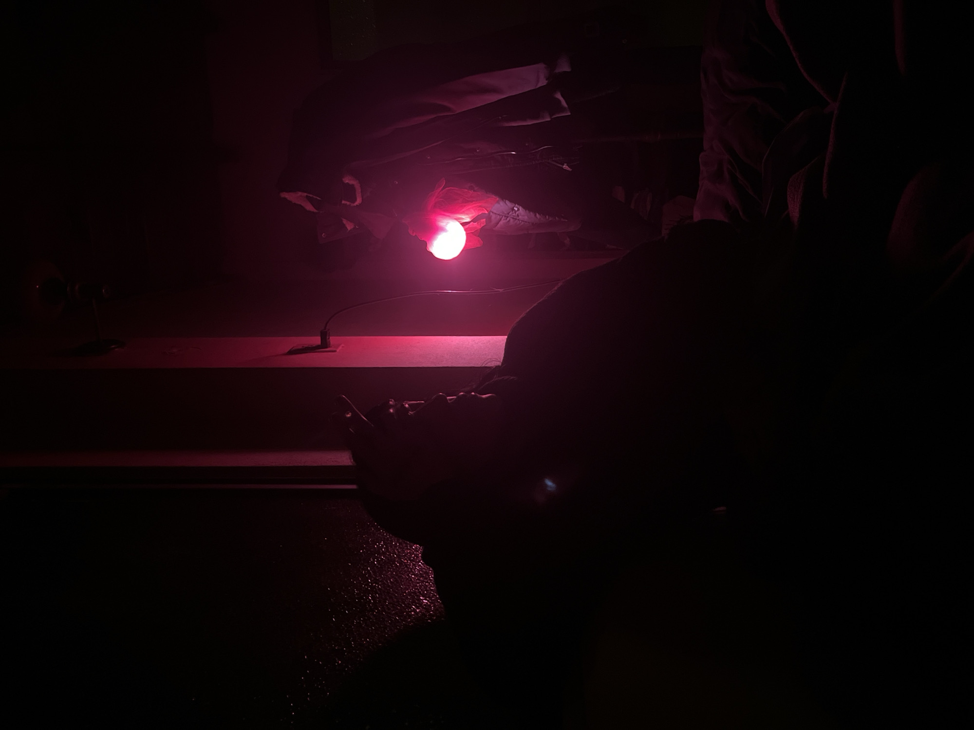 This dimly lit image portrays an indoor scene dominated by a strong red hue emanating from a central light source. A bright, glowing red light bulb or lamp is positioned in the upper center, illuminating some dark fabric or clothing items above it. A black cord extends from this light.
In the lower-center of the frame, a person is visible, largely in silhouette, with their head bowed or resting on a surface. Their posture suggests they are kneeling, crouching, or leaning forward, and their features are obscured by shadow. Below and to the left of this figure, a reflective, textured surface, possibly wet with water or another liquid, catches the red light, creating a sparkling effect.
To the far left, a small, dark object, resembling a table lamp or a piece of equipment on a stand, is visible on a flat surface. Another dark, indistinct shape, possibly part of a person or furniture, is present in the upper right portion of the image. The environment appears to be a darkened room, likely at night, with the artificial red light providing the sole illumination. The overall mood is mysterious and contemplative. No discernible text is visible in the image.