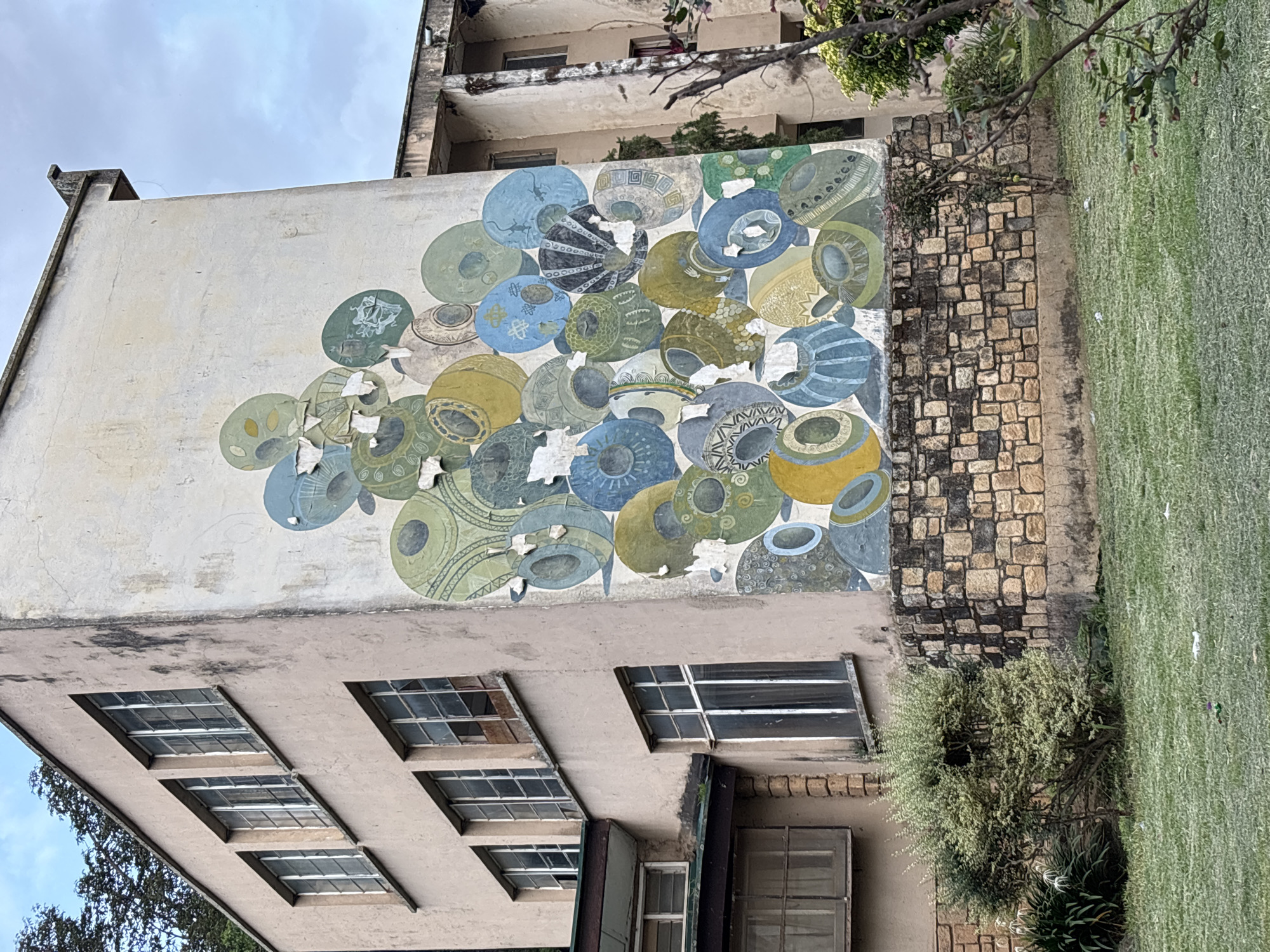 A side view of a weathered, multi-story building in Jos, Nigeria, is dominated by a large, faded mural on its exterior wall. The mural, composed of circular shapes in shades of blue, green, yellow, and grey, depicts what appears to be a collection of stylized pots or vessels. Some areas of the mural show peeling paint. The building itself is a light beige color, with multiple rows of windows featuring metal frames. The lower portion of the building is adorned with a stone foundation. In front of the building, a patch of green lawn is visible, with a small, leafy bush in the foreground. The sky above is overcast.