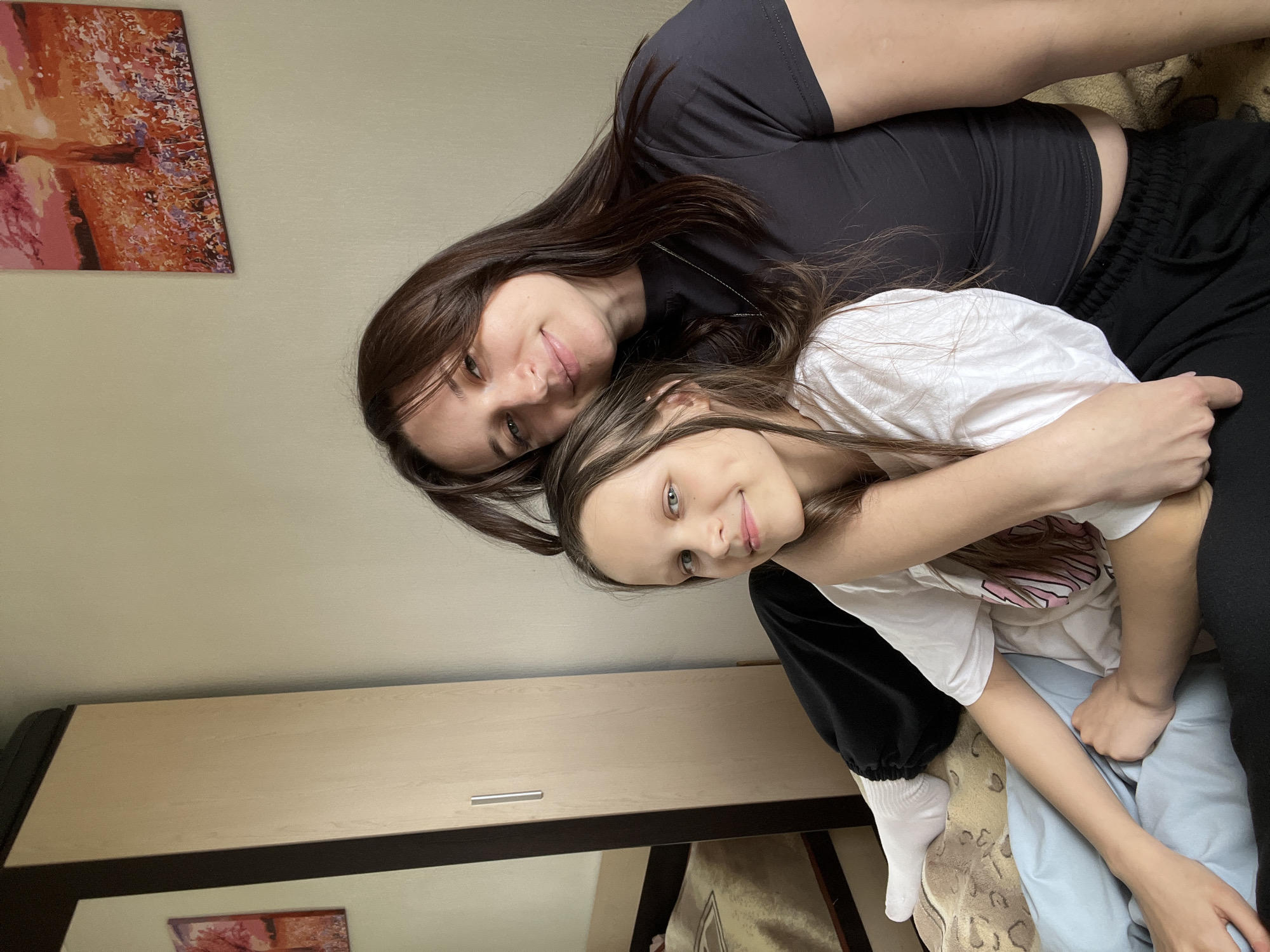A woman and a girl are embracing indoors in Usol'tseva, Russia. The woman has dark brown hair, is wearing a black crop top and black sweatpants. The girl has light brown hair and is wearing a white t-shirt, black sweatpants, and one white sock. They are sitting on a bed covered with a patterned bedspread and a blue blanket. Behind them is a light tan wall with a painting on it and a light brown dresser with a dark brown trim. The woman is lying on top of the girl and they are both smiling at the camera.