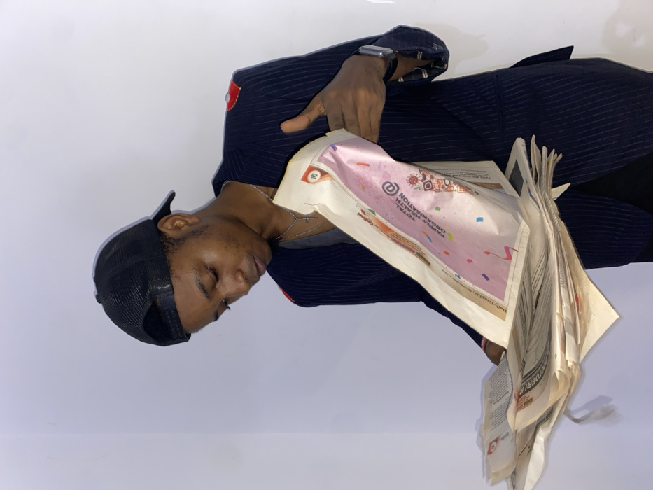 In Joma, Ghana, a young man is captured in a half-body shot against a plain, light grey or white background, suggesting an indoor or studio setting. He wears a dark, possibly navy, pinstriped suit jacket and a black trucker-style cap. A silver chain necklace with a small pendant hangs around his neck, and a dark-strapped wristwatch is visible on his left wrist.
The man's eyes are closed or heavily lidded, lending him a pensive, calm, or introspective expression. He holds a stack of newspapers against his chest with both hands. One newspaper page, prominently visible, features a colorful design with pink and purple hues, and appears to be an advertisement or special section. On this page, the text "TOTAL HEALTH ORGANISATION" is clearly legible, accompanied by a circular logo. Below it, the words "FEDERAL ORGANISATION" can be partially seen. A small box containing the number "29" is visible near the top left of this page, and the newspaper's masthead "Daily Graphic" is visible lower down. Other newspaper pages are stacked below this one, showing typical black-and-white print, but with no decipherable text. The lighting is soft and even, highlighting the subject's features and the details of the newspapers.