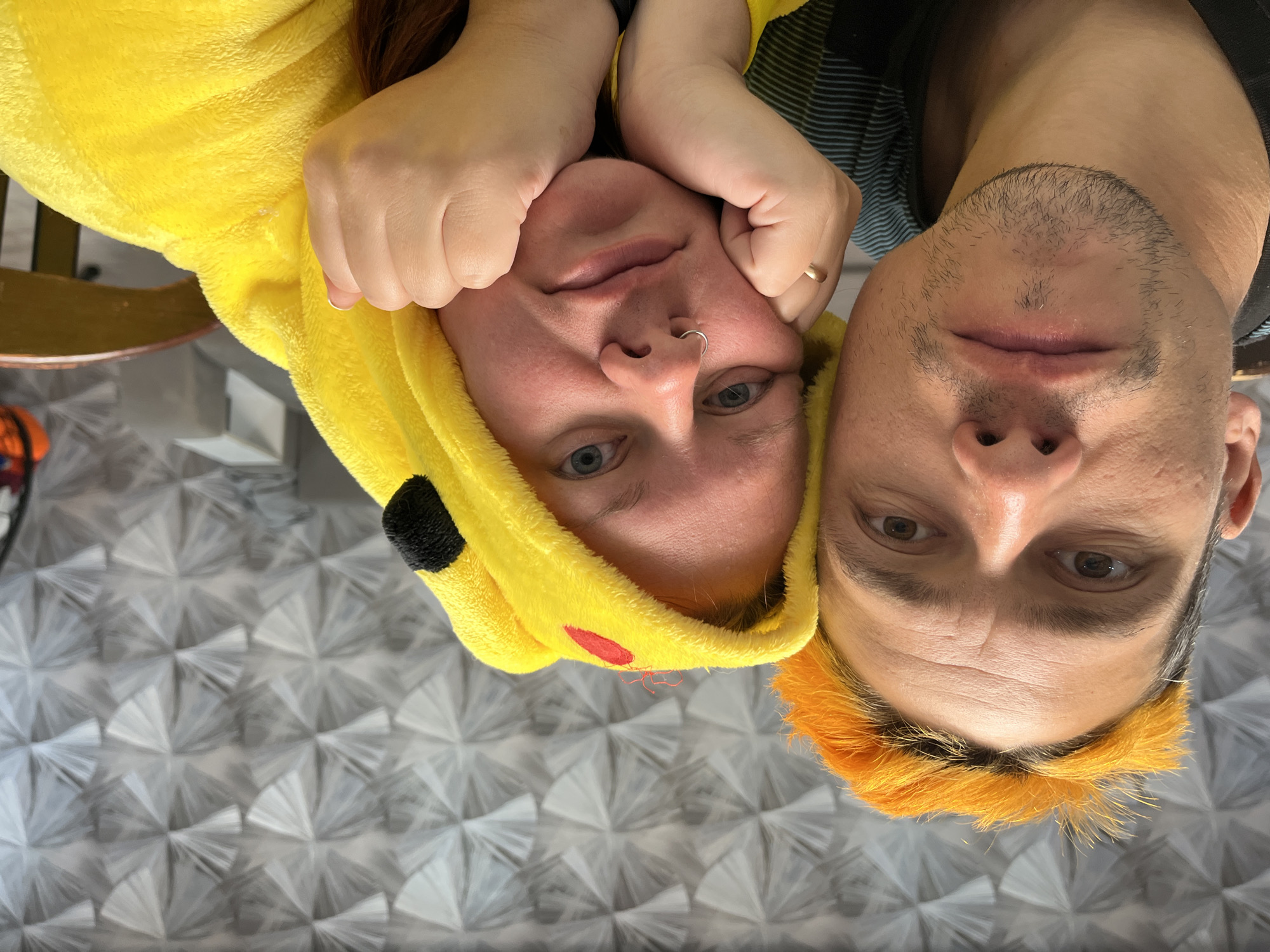 An inverted close-up selfie features two individuals, a woman and a man, positioned upside down against a patterned background.
The woman, on the left, is wearing a bright yellow, fluffy hooded garment that resembles a Pikachu costume, complete with black ear tips and a red cheek detail on the hood. Her reddish-brown hair is visible beneath the hood. She has a silver nose ring and blue eyes, and her hands are raised, gently cupping her cheeks. Her expression is calm, with a slight hint of a smile as she looks directly at the camera.
The man, on the right, has short hair with a distinct bright orange dye at the front, creating a striking contrast. He has light stubble on his face and wears a dark top with visible blue stripes on the shoulder. A gold-toned ring is visible on the ring finger of his left hand, which is positioned near the woman's head. His eyes are brown, and he also looks directly at the camera with a neutral expression.
The background is an indoor surface, possibly a floor or wall, featuring a repetitive geometric pattern of interconnected grey and white fan-like or radial shapes that appear to have a reflective or textured quality. A small portion of a wooden chair or table leg is visible in the upper left corner. The casual attire, including the playful costume, suggests a relaxed and informal moment. No discernible text is visible in the image.