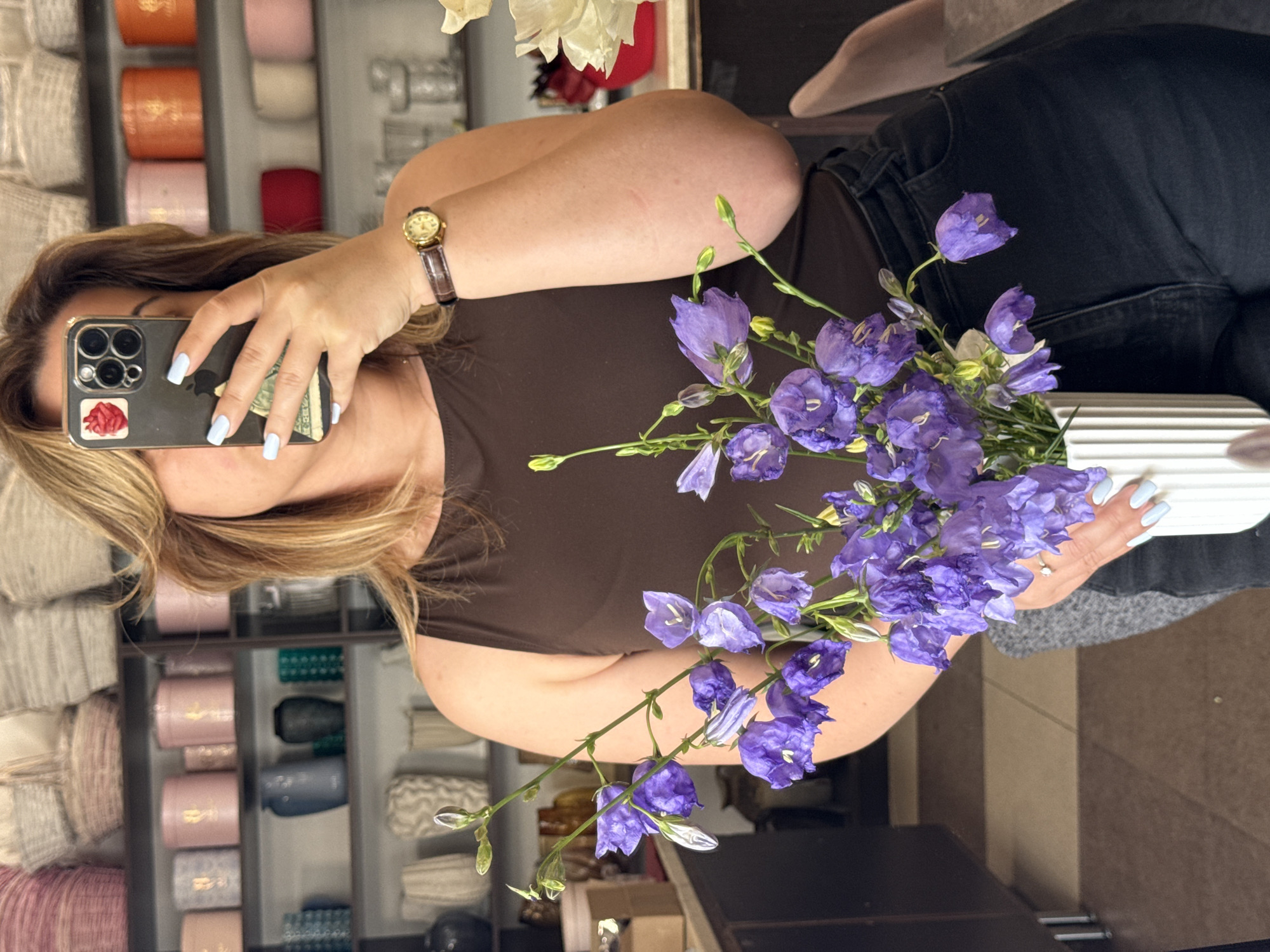 The image shows a woman taking a selfie in a retail store, possibly a flower shop or a home goods store. She is holding up her phone, which has a red sticker on the back. The woman has blonde hair and is wearing a brown top and black jeans. She is also wearing a watch with a brown band on her left wrist and has light blue nail polish. A bouquet of purple flowers is in the foreground, obscuring part of her face and phone. In the background, shelves are filled with various items, including rolls of fabric or yarn in pastel colors and some decorative items. The lighting suggests it is daytime.