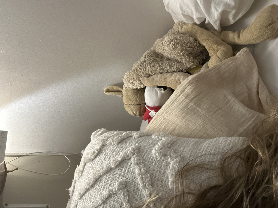 A close-up depicts a cluttered bed area. A plush sheep rests atop a white stuffed egg wearing a red scarf. Beige pillows and blankets surround the toys, partially obscuring them. A textured white pillow with a geometric pattern is in the foreground. Loose strands of brown hair are also visible on the right. A white wall and a glimpse of a lamp and its cord can be seen in the background.
