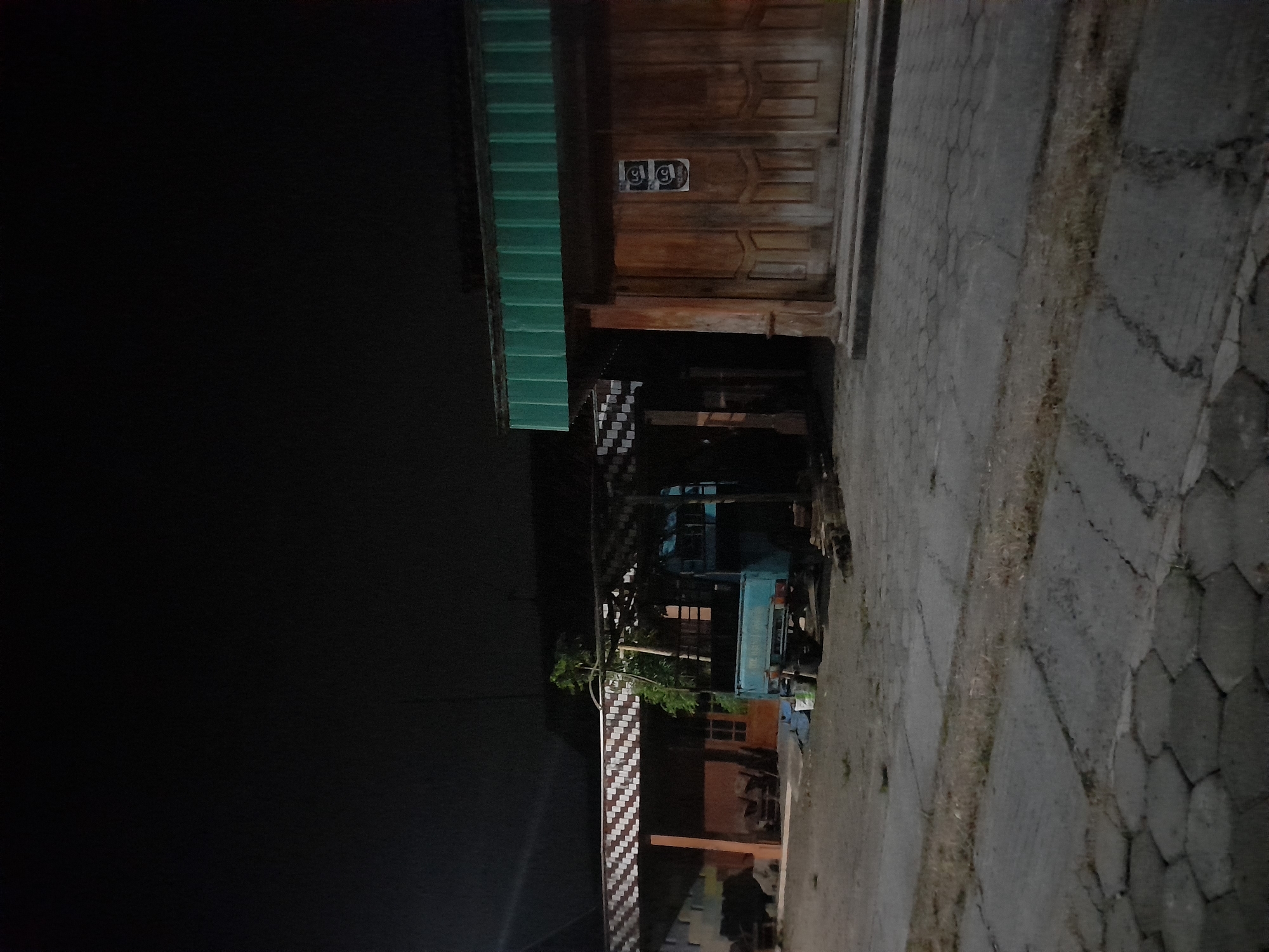 The image depicts a street scene at night in Pulokulon, Indonesia. The setting appears to be residential or commercial, with a building featuring a wooden door with carved panels visible on the right side. Two stickers with the letters "gg" are affixed to the door. Adjacent to the building is a green corrugated roof extending over a portion of the scene. A light blue truck is parked in front of the building, partially illuminated by ambient light. The ground is paved with interlocking stones and some dirt or grass. The overall impression is one of a quiet, dimly lit evening. There are no people visible in the frame. The time of day is clearly night, and the weather is not discernible.