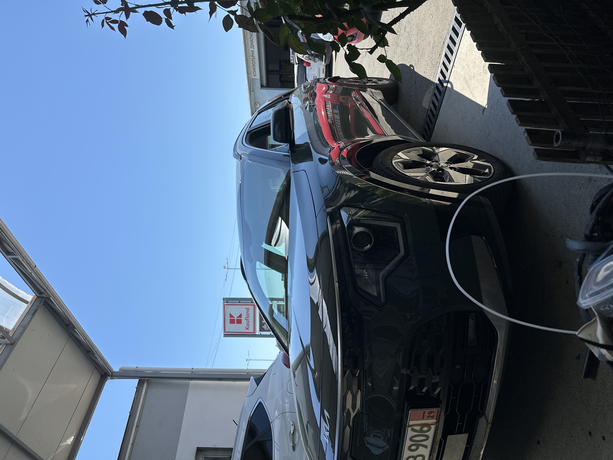 The image shows a black car parked outdoors under a covered structure. A red sign with a white "K" and the word "Kaufland" is visible in the background, suggesting the location is near a Kaufland supermarket. To the right, the building has text that reads "КОМПЮТЪРНА ДИАГНОСТИКА," indicating a computer diagnostics service. The sky is clear and blue. A plant with dark leaves is growing in the foreground on the right. The car's license plate is partially visible, reading "B 906" and the number "29".