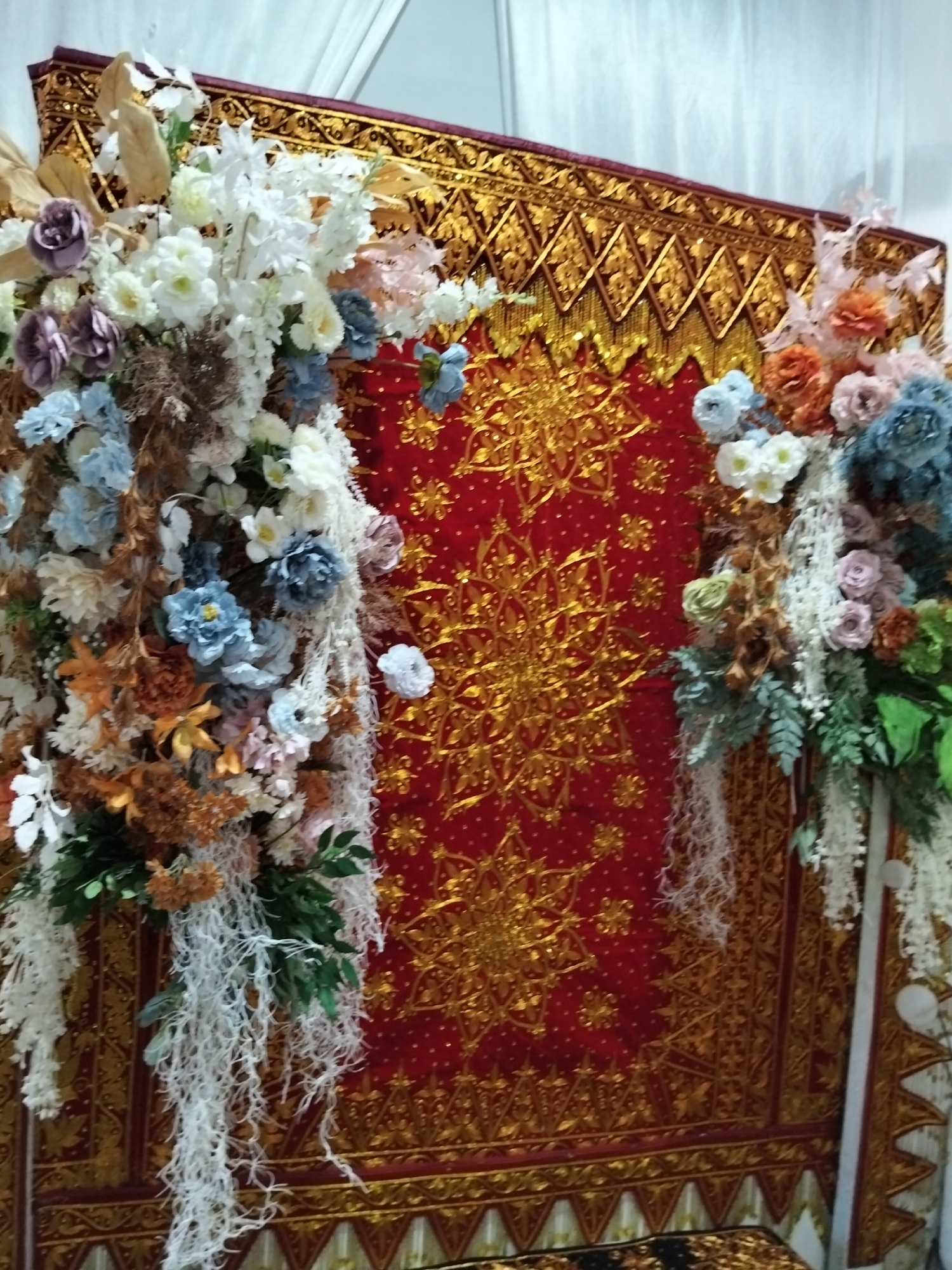 The image depicts a ceremonial backdrop featuring a red fabric with intricate gold patterns. This backdrop is adorned with elaborate floral arrangements on both the left and right sides. The floral arrangements consist of a variety of artificial flowers in colors such as white, blue, pink, and orange, along with dried elements and greenery. Draped white strings or netting hang from these arrangements. The backdrop itself has a rich, golden trim along its top and bottom edges, with geometric designs. The setting appears to be indoors, possibly for an event like a wedding or formal ceremony, given the decorative nature of the scene. The lighting suggests an indoor environment, possibly with some ambient light. The location in Pulonga, Indonesia is inferred from the provided context.