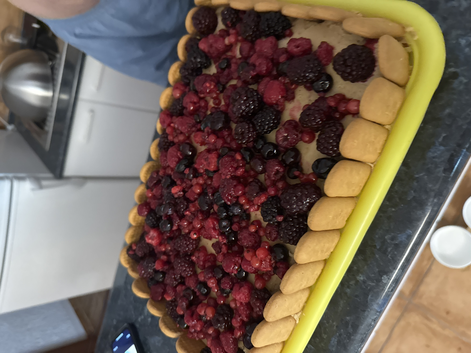 An overhead shot captures a rectangular, freshly made cake, presented on a bright yellow plastic serving tray. The cake's base is a pale yellow filling, topped generously with a vibrant mix of red and dark berries, including raspberries, blackberries, and red currants. The perimeter of the cake is adorned with a decorative border of light brown ladyfinger biscuits. The cake and tray rest on a dark, speckled countertop. In the background, a stainless steel appliance, possibly a kettle or pot, is visible on a stovetop. To the left of the cake, a smartphone is placed on the countertop, displaying a bright screen. The overall environment appears to be a domestic kitchen setting. The location is identified as Zankovići, Montenegro.