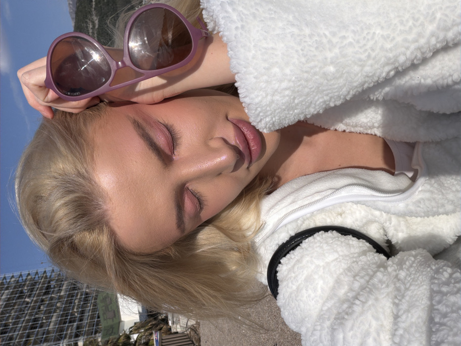 A close-up, outdoor selfie depicts a fair-skinned individual with blonde hair, closed eyes, and a slight smile, adorned with pink-toned eyeshadow and lip color. The person wears a white, textured sherpa-style zip-up jacket over a white garment, with a black strap visible over the left shoulder. The right hand holds a pair of large, purple-framed sunglasses with dark, reflective lenses (with "GREG CH..." faintly visible on the right arm) against the right temple. The background features a clear blue sky. To the left, a multi-story, unfinished concrete building with extensive scaffolding is visible, situated above green foliage and lighter-colored structures. The ground in the foreground appears to be light-colored and textured, consistent with a beach or promenade. Distant mountains are present on the right horizon. This scene is located in a coastal area of Budva, Montenegro.