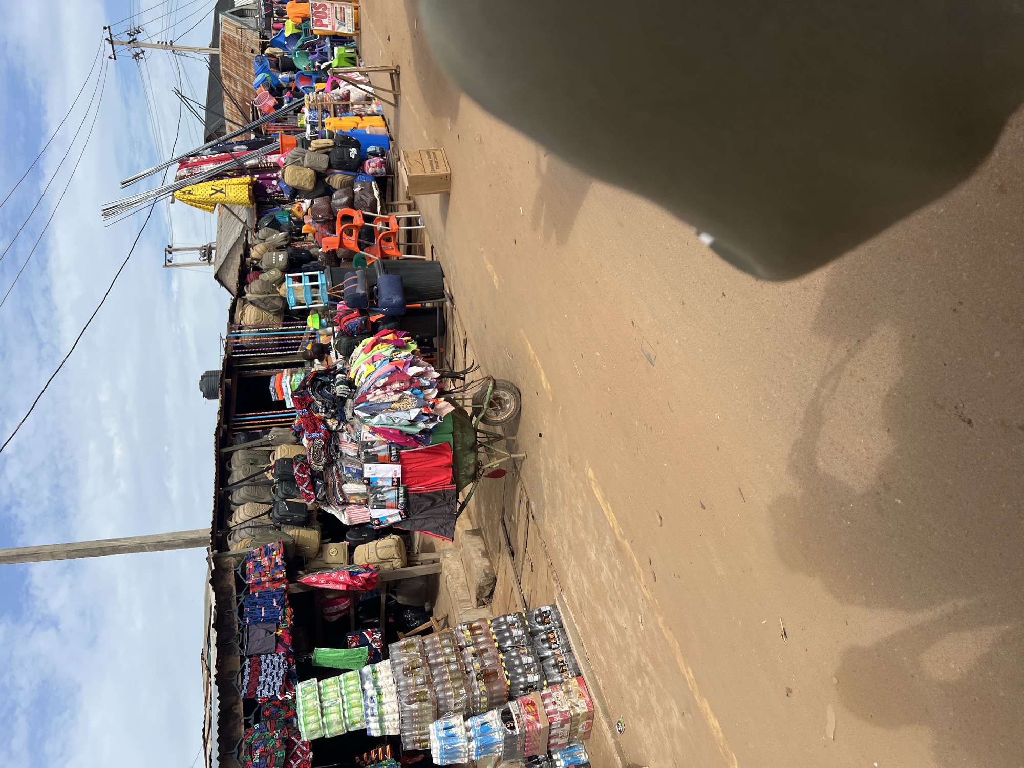 The image depicts a busy street market in Markurdi, Nigeria. Stalls line the street displaying various goods, including clothing, bags, and beverages. A cart laden with merchandise is visible, and there are stacks of packaged drinks on the ground. The environment suggests an active commercial area.