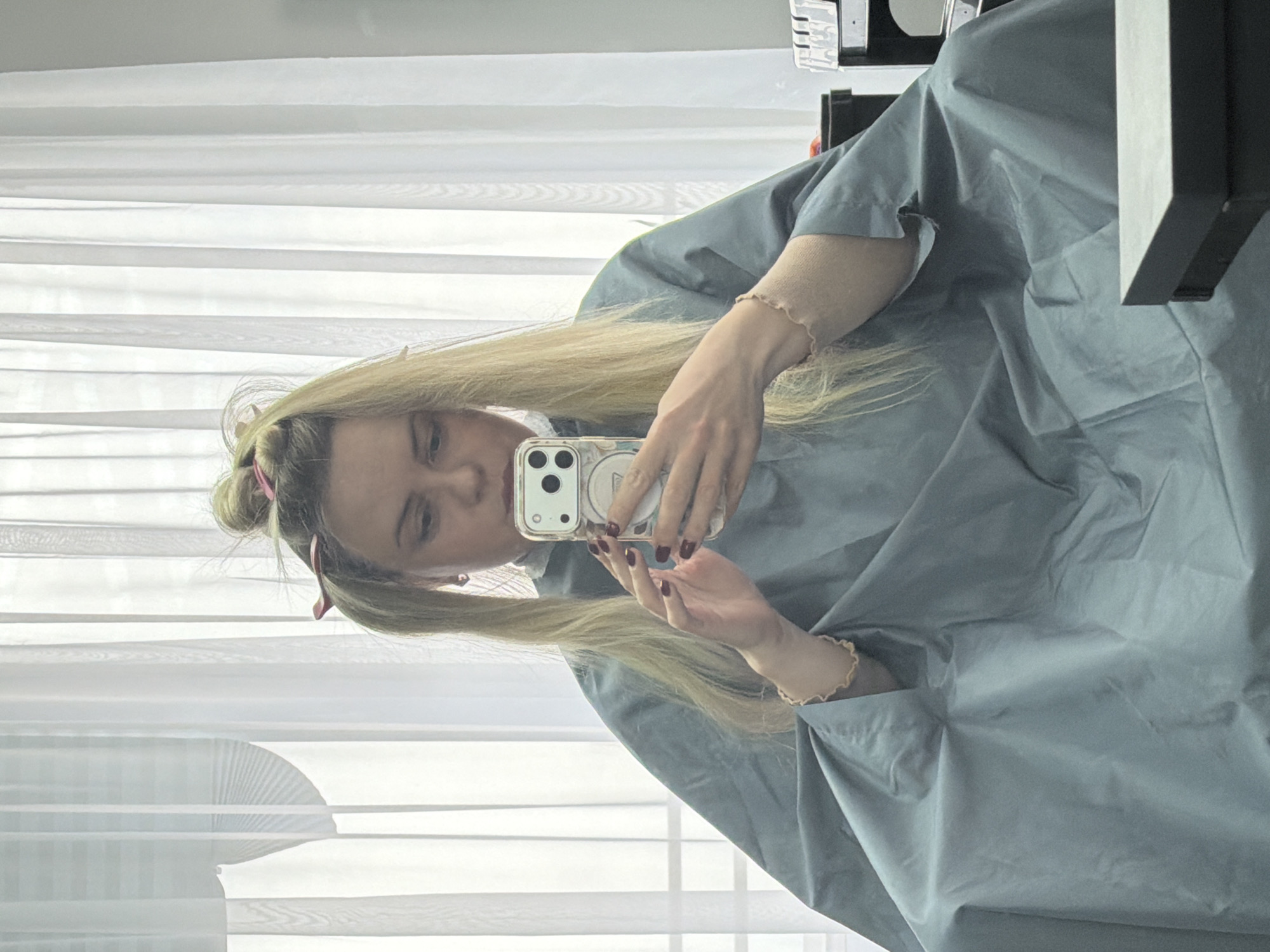 A woman with long blonde hair is taking a selfie with her smartphone in what appears to be a beauty salon. She is wearing a light blue smock or cape. Her hair is pulled back with pink clips. She has a bracelet on her left wrist and red nail polish on her fingers. The background features white blinds or curtains, and there is some salon equipment visible to the right of the frame. The location cannot be confirmed, as the city is unknown.