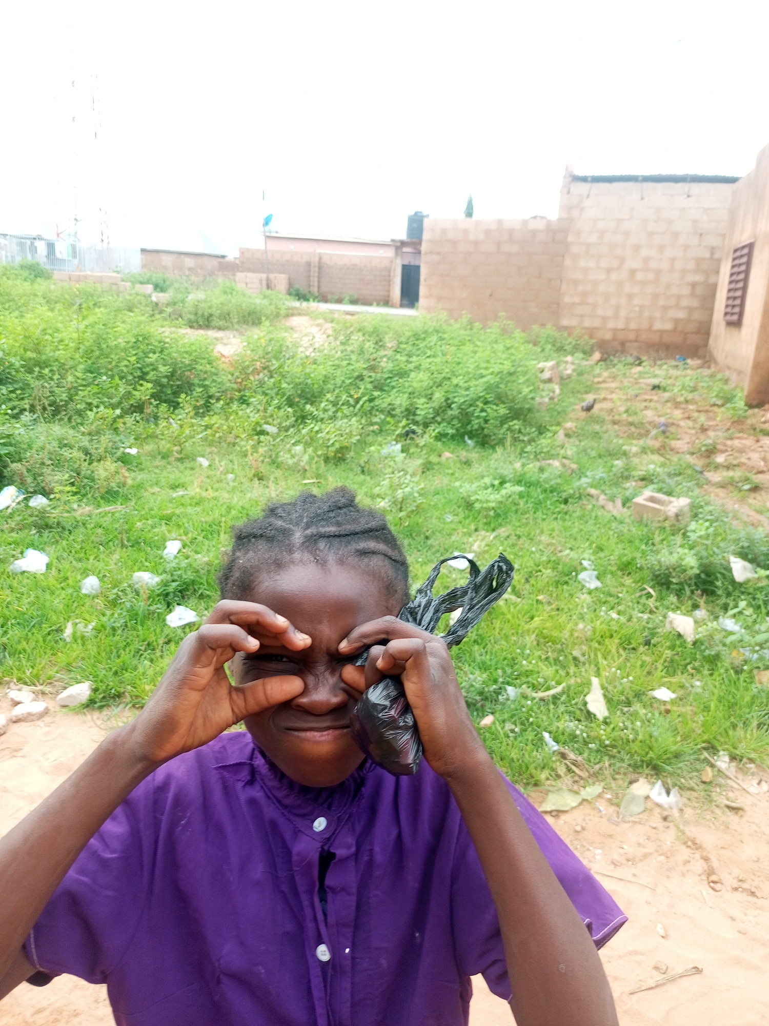 A young Black individual, wearing a purple button-down shirt, is looking directly at the camera. Their hands are positioned around their eyes, with a black plastic bag held near their face. The background consists of overgrown green grass, scattered white debris, and partially constructed buildings. The scene appears to be in an outdoor, somewhat unkempt environment, possibly on the outskirts of a residential area in Tempure, Nigeria. The lighting suggests it is daytime.