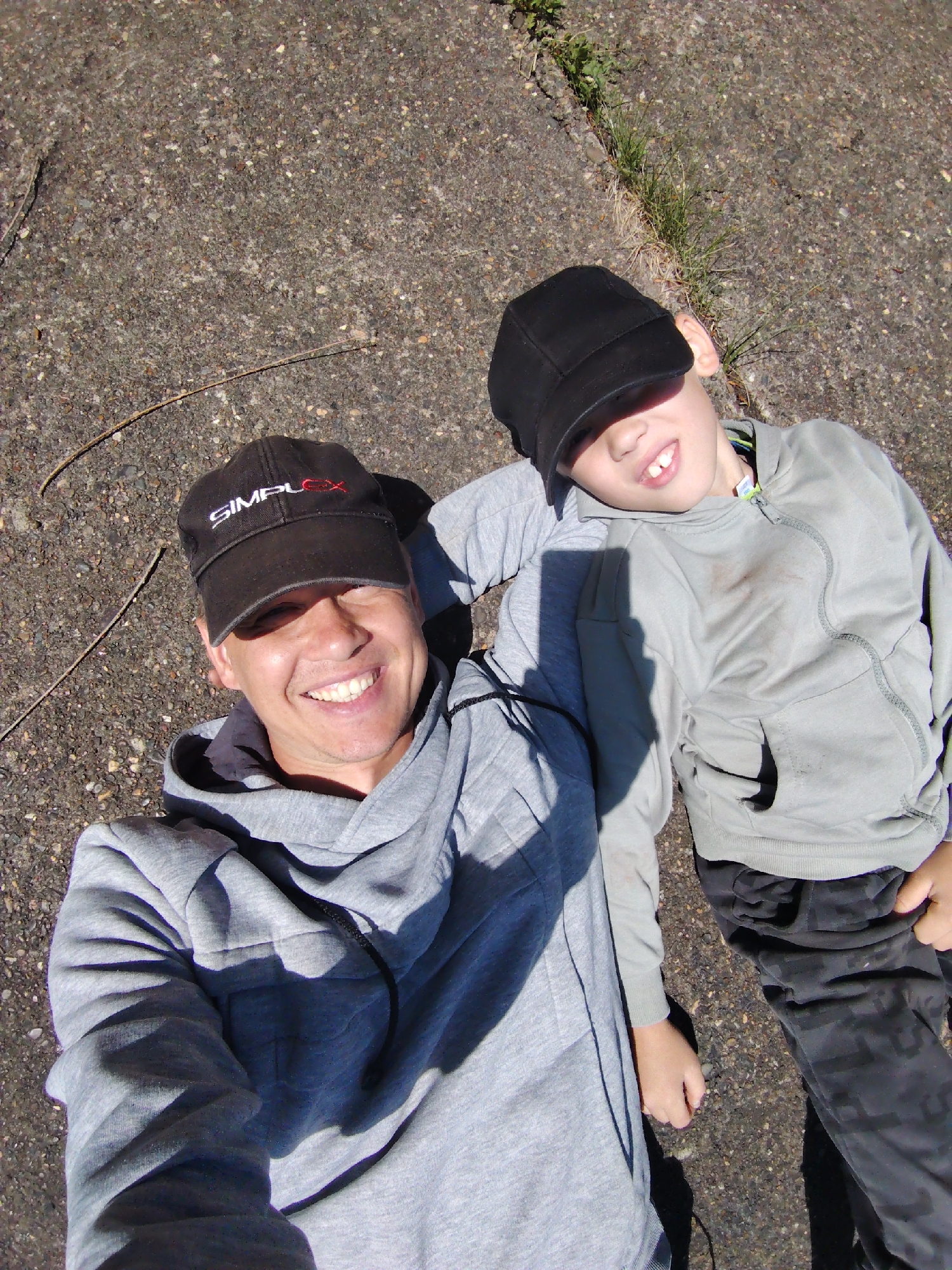 A medium-angle, eye-level shot shows an adult man and a child lying on a paved surface. The man, positioned on the left side of the frame, is wearing a gray hooded sweatshirt and a black baseball cap with white and red lettering, and is smiling broadly. To his right, a child is lying down with their head resting on the man's arm. The child wears a gray hooded sweatshirt, black pants with a camouflage pattern, and a black baseball cap obscuring their eyes.
The surface they are lying on appears to be rough asphalt or pavement. A thin, dry twig lies near the man. A narrow crack in the pavement, filled with grass, is visible near the top right of the frame. The lighting suggests an outdoor setting, likely during the daytime.
There is no verifiable information in the image that indicates the location to be Chaikovsky, Russia.