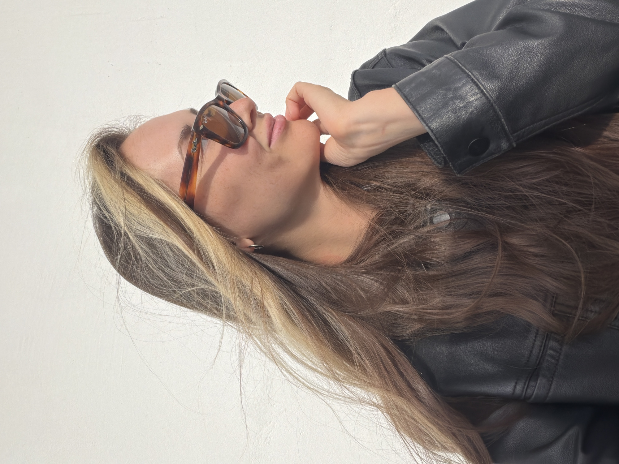 An outdoor medium shot shows a woman with long, layered brown and blonde hair against a white wall. She is wearing a black leather jacket and brown-rimmed sunglasses. Her right hand is resting on her chin, and she is looking to the right. The lighting suggests it is daytime, with a slight shadow cast on the wall behind her. There are no other people or significant objects visible in the frame. The location is identified as Khimki, Russia.