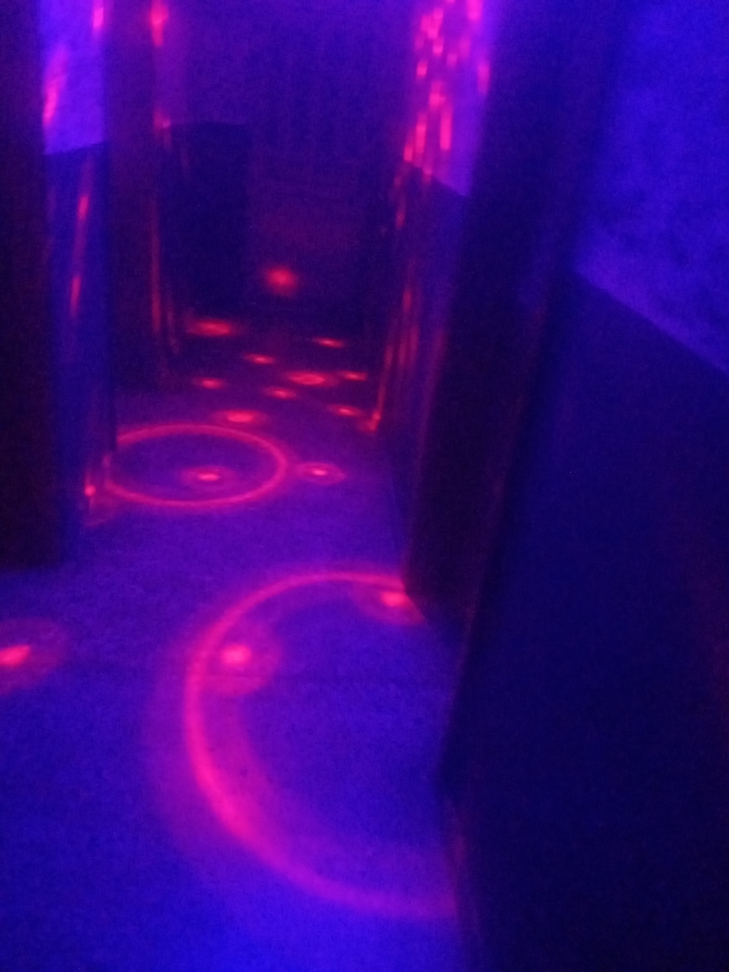 The image depicts an indoor environment illuminated by purple and pink lighting, suggesting a place like a laser tag arena or a similar entertainment venue. The floor is a dark, possibly carpeted surface, marked with glowing pink circles of light. These circles appear to be projected onto the floor, with some circular patterns also visible in the background and on the walls. The overall ambiance is dark and atmospheric, with the lighting creating a sense of depth and mystery. The architecture suggests narrow passages or walls, adding to the enclosed feel of the space. The image does not contain any individuals or specific objects that would indicate a particular location within Sabon Gari Nassarawa, Nigeria. The scene is devoid of people and any recognizable landmarks or cultural elements.