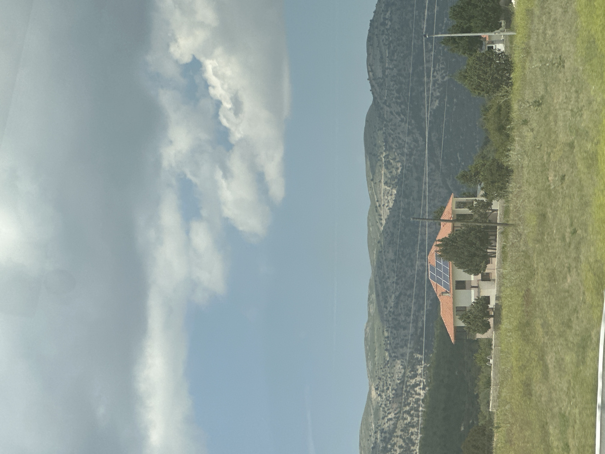 The image shows a rural landscape in Korkuteli, Turkey, with a house prominently featured in the midground. The house has a red tile roof and solar panels installed on it. It is surrounded by greenery, including trees and bushes. In the background, there are rolling hills covered in sparse vegetation and trees, with a clear sky above. The sky is partly cloudy, with a mix of light and dark clouds. Power lines are visible stretching across the scene, indicating the presence of infrastructure. The foreground consists of a grassy field. There are no people or animals visible in the image.
