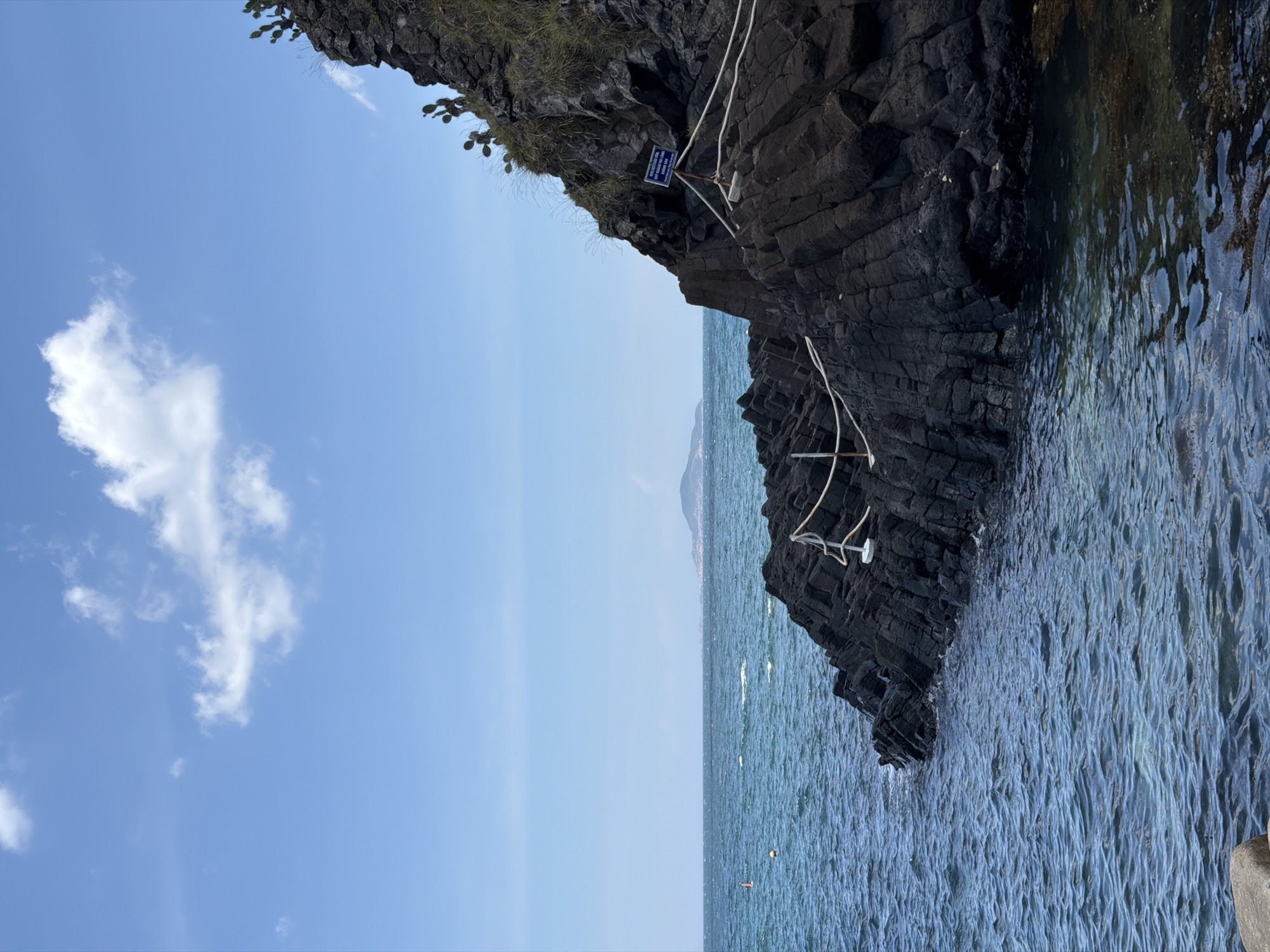 The image depicts a coastal scene in Phú Lương, Vietnam. In the foreground, dark, layered volcanic rocks form a rocky shoreline. The sea is a vibrant blue, with gentle waves lapping against the rocks. A rope barrier, supported by white posts, guides visitors along the edge of the rocks. In the distance, the vast expanse of the sea stretches to the horizon, meeting a clear blue sky dotted with a few fluffy white clouds. A small, hazy island is visible in the far distance. The overall impression is one of natural beauty and tranquility, with hints of human presence through the rope barrier and a small buoy in the water.