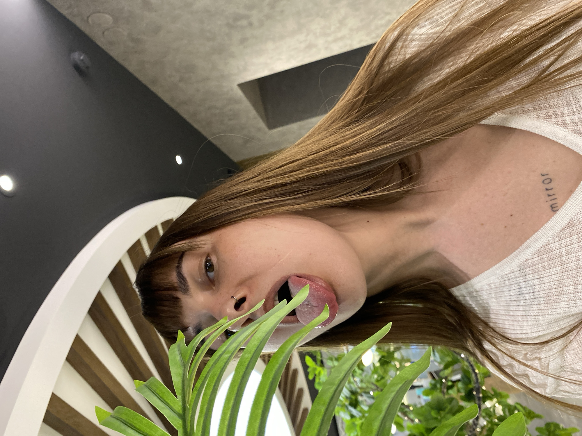 A young woman with light brown hair and bangs sticks her tongue out towards a plant in front of her. She has a nose ring and a tattoo on her collarbone. The background includes a black wall, a patterned decorative element, and a blurred image of plants and a window. There is no indication of this location being in Votkinsk, Russia.