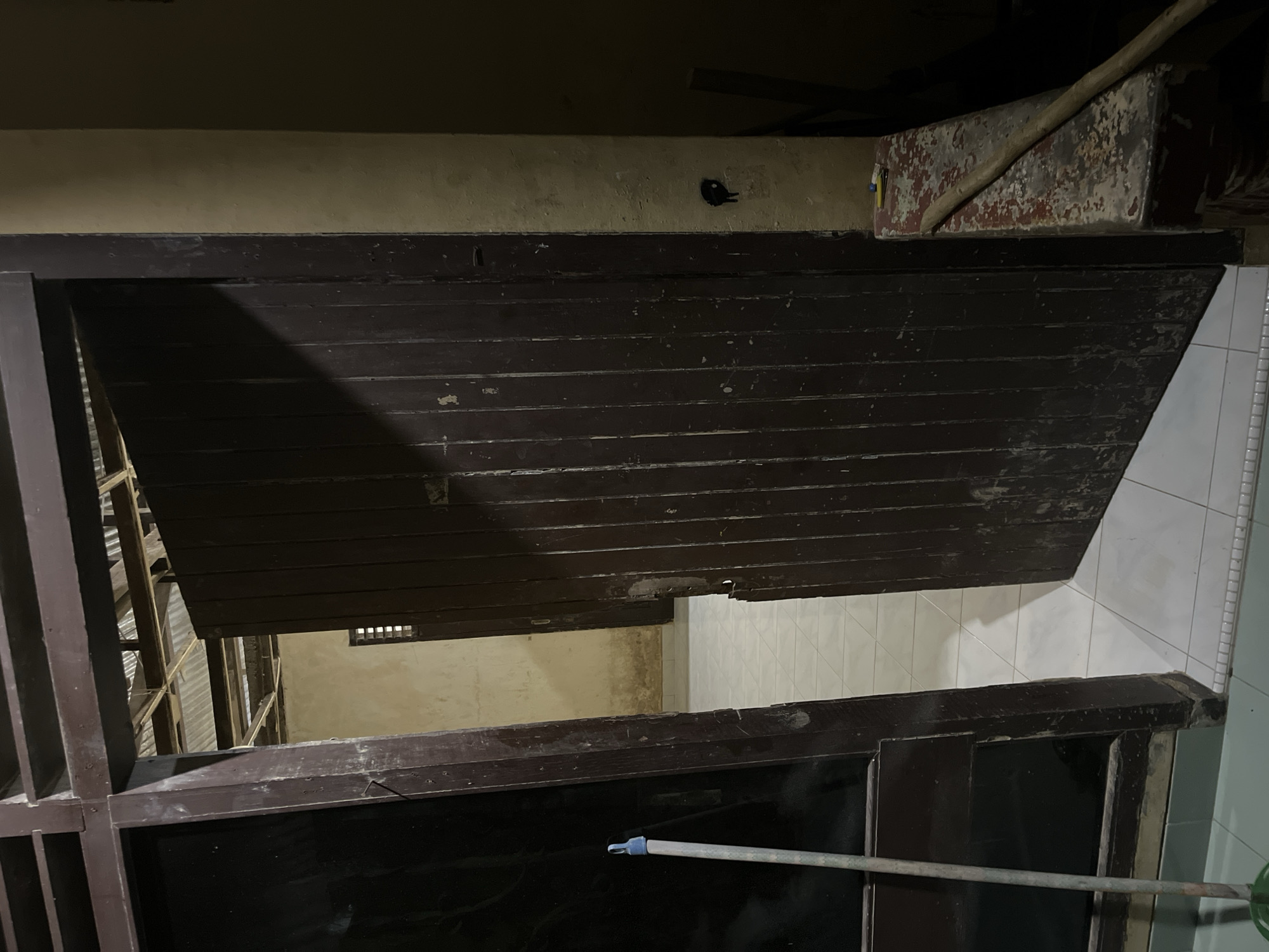 The image shows an interior view of what appears to be a hallway or entryway in Kota Kuala Simpang, Indonesia. In the foreground, a dark brown wooden door is open, revealing a glimpse of another room beyond. To the left, a dark, reflective surface, possibly another door or a large window, is visible. A light-colored broom with a blue handle stands upright in the lower left corner. The floor is tiled with white squares. The walls are a muted, textured yellow. The ceiling appears to be constructed of corrugated metal sheets supported by wooden beams. In the background, through the open doorway, another dark wooden door can be seen. The overall impression is of a simple, functional interior with signs of wear and tear.
