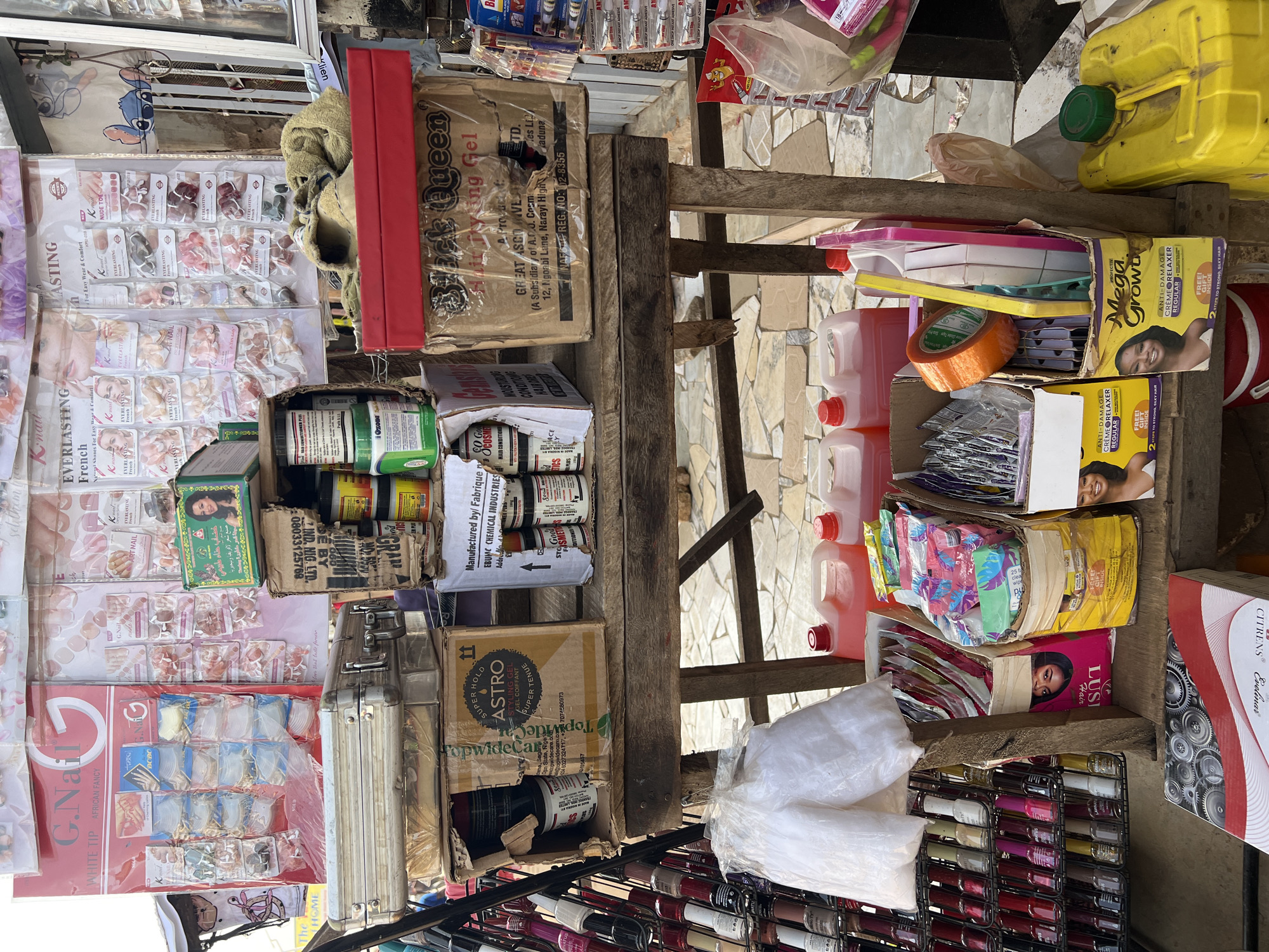 The image captures a vibrant street-side stall in Bukuru, Nigeria, showcasing an array of beauty and hair care products. The stall is a colorful medley of merchandise stacked on wooden shelves and displayed on a metal rack. Prominently featured are various hair products, including gels and relaxers, with brands like "Black Queen" and "Mega Growth" visible. A selection of nail art supplies, such as artificial nails and nail polish, are also displayed. Interspersed among these are containers of liquid, possibly cleaning supplies or beauty treatments, and rolls of tape. A vintage metal suitcase sits on the top shelf, adding a touch of nostalgia. The overall impression is that of a bustling marketplace, typical of many Nigerian cities, where local entrepreneurs offer a wide range of goods to cater to consumer needs.