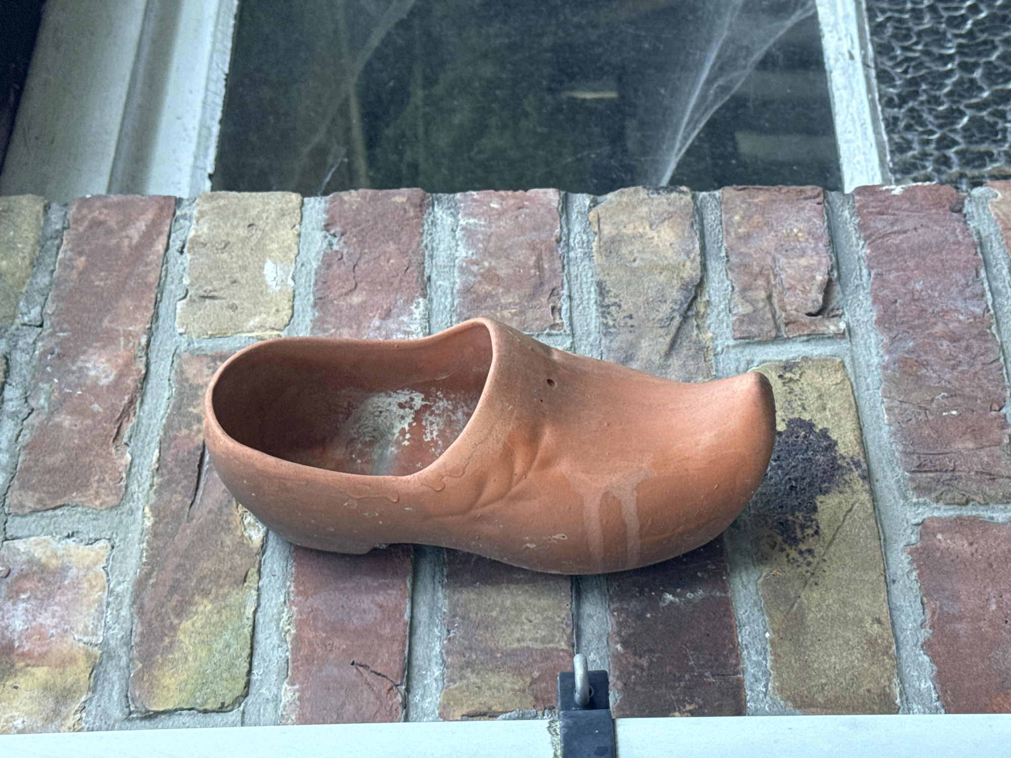 The image shows a terracotta clog, likely a planter, mounted on a brick wall. The clog is oriented vertically, with its opening facing upwards and to the left. It appears to be empty and shows signs of weathering, with some white residue and streaks on its surface. The brick wall is constructed with irregular, reddish-brown and yellow bricks, mortared with light grey grout. To the right of the clog, a window pane is visible, partially obscured by shadows and what appears to be cobwebs. The window frame is light colored, possibly white or pale blue. A black metal latch is attached to the left side of the wall. The overall scene suggests an outdoor setting, possibly a garden or the exterior of a building. There are no people or animals visible in the image. The image does not provide specific geographical markers to pinpoint a location within Bruges, Belgium.