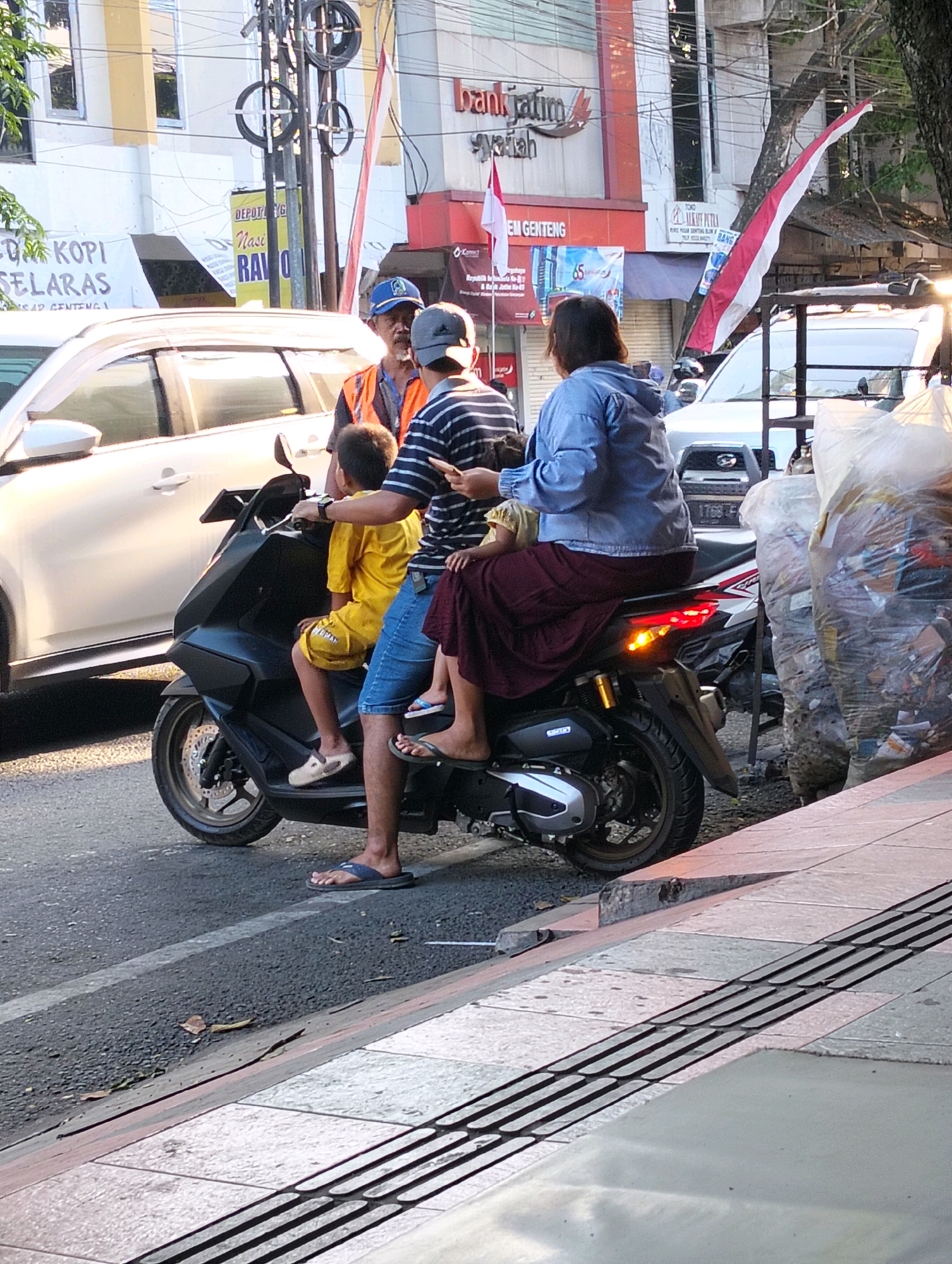 The image shows a street scene in Genteng, Indonesia, with a group of people on a scooter. In the foreground, a black scooter is parked on the side of the road. A young boy in a yellow shirt and shorts sits in front of a man wearing a blue and white striped shirt and blue denim shorts. Behind the man is a woman wearing a blue jacket and a maroon skirt. An older man in an orange vest and blue cap stands near the scooter, facing the people on it. The street is paved with asphalt, and a sidewalk with tactile paving is visible to the right. In the background, there are buildings, including one with "bank jatim syariah" signage, and a white car. The lighting suggests it is daytime, possibly late afternoon, with sunlight casting shadows. Several large plastic bags filled with refuse are also visible on the right side of the image.