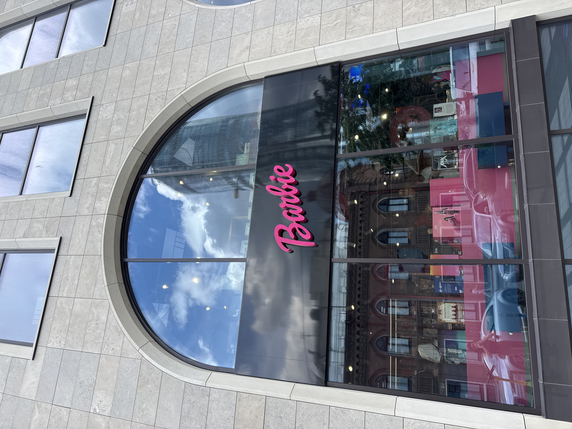 The image shows the exterior of a Barbie store in Yekaterinburg, Russia, located on Malysheva Street. The store features a large arched window with the "Barbie" logo in pink lettering. Reflected in the window are a modern skyscraper and the facade of an older building across the street. Inside the store, visible through the window, are displays of Barbie merchandise, including dolls and pink accessories, as well as a light blue car and a pink car. Two women are visible inside the store, one of whom is taking a photo. The building's facade is clad in light-colored stone tiles.