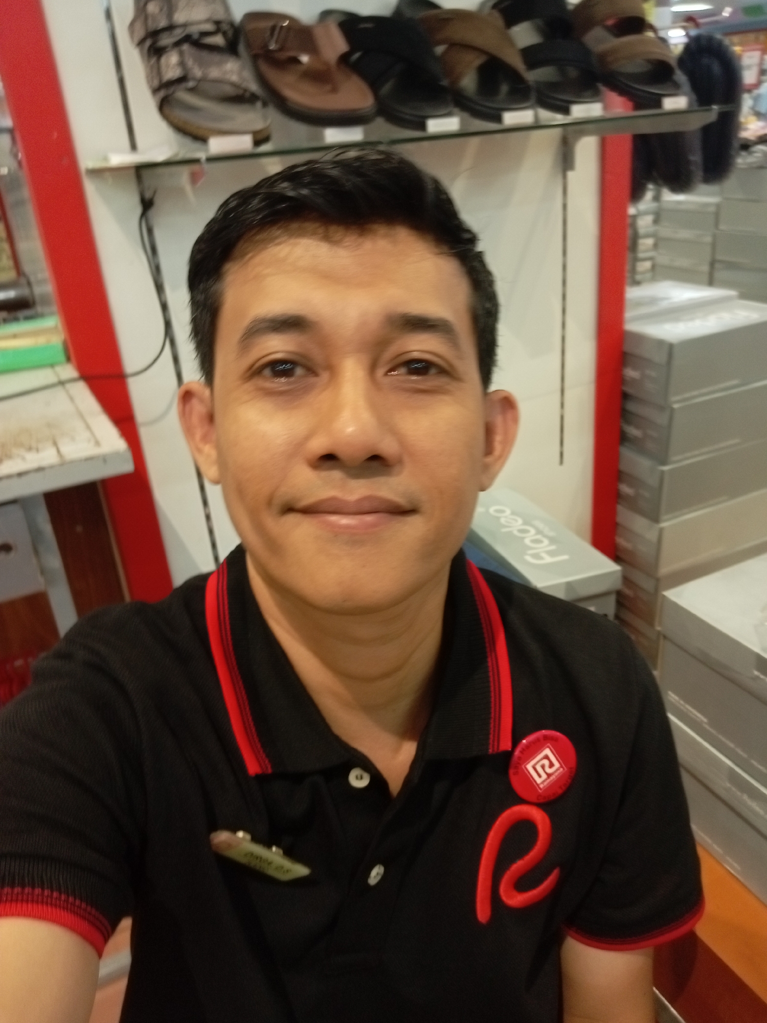 In Batam, Indonesia, a man is captured in a selfie, wearing a black polo shirt with red trim and red embroidery. He has a name tag clipped to the left side of his shirt, and a circular red badge with a logo on the right. The background shows shelves stocked with shoes and shoe boxes, suggesting the setting is a retail store. The man's expression is neutral, and he is looking directly at the camera. The lighting indicates an indoor environment.