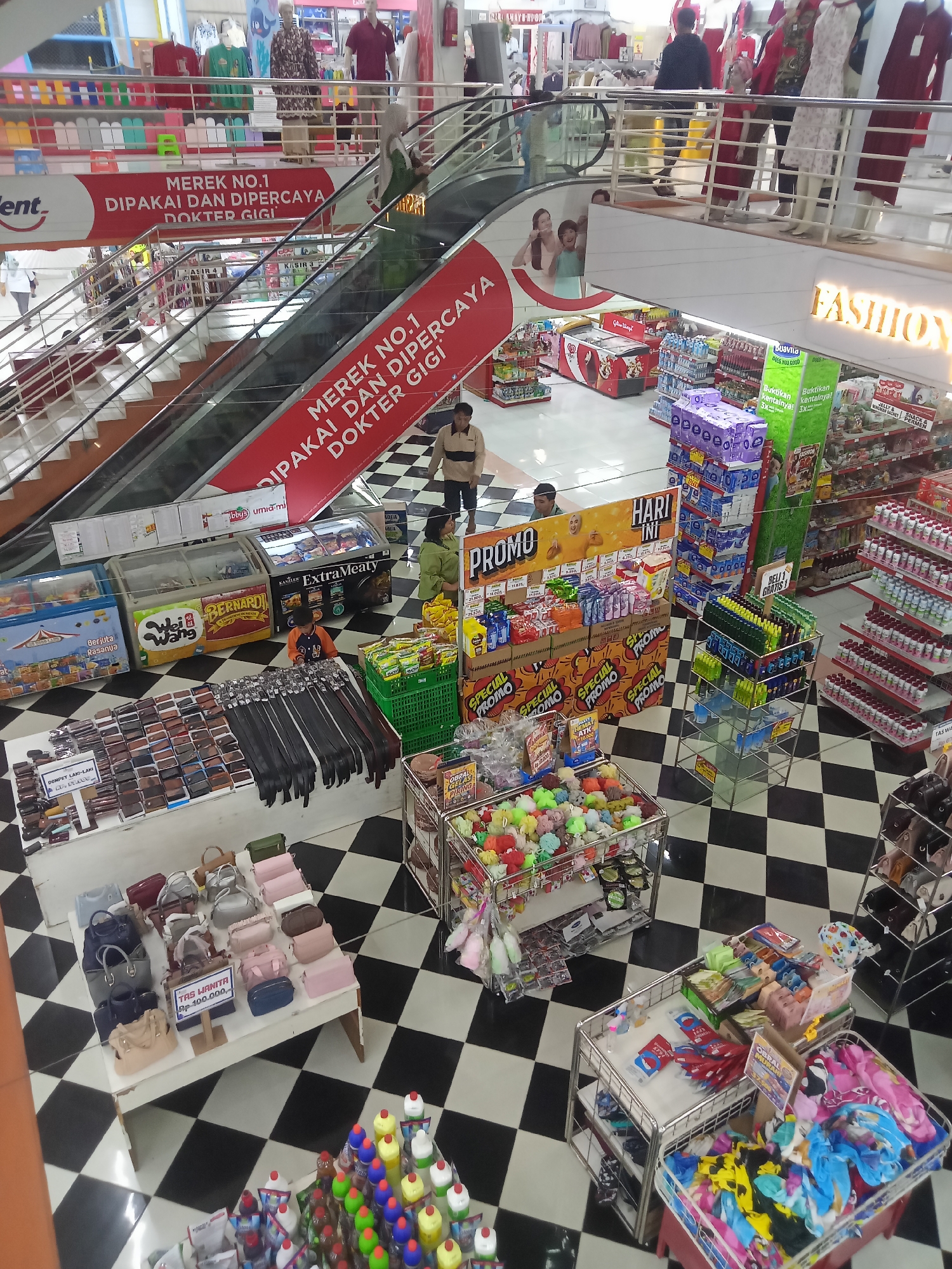 This image depicts the interior of a busy department store, likely in Boyolali, Indonesia, characterized by its black and white checkered flooring. Several people are visible, including shoppers browsing merchandise and employees attending to displays. The store features multiple levels, with escalators facilitating movement between them. Merchandise is displayed prominently on shelves and tables, including clothing on mannequins, bags, wallets, cleaning supplies, and various packaged goods. A large red banner with white text reading "MEREK NO.1 DIPAKAI DAN DIPERCAYA DOKTER GIGI" (Brand No. 1 Used and Trusted by Dentists) is prominently displayed on an escalator. Another sign reads "FASHION". Promotional displays with "PROMO" signage are also evident, suggesting ongoing sales. The overall atmosphere appears to be a typical retail environment with a diverse range of products available.