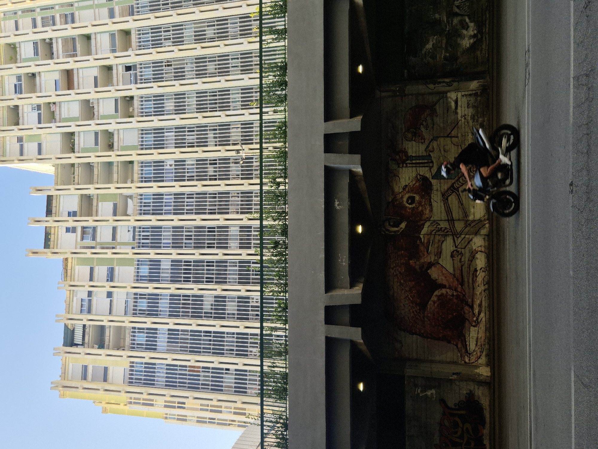 The image depicts a street scene in Taranto, Italy, with a cyclist riding a motorcycle under a viaduct. The viaduct's underside is adorned with graffiti, featuring a large depiction of a rabbit and other artistic elements. Above the viaduct, a modern building with a distinctive facade of vertical elements and numerous windows rises against a clear blue sky. A green railing with small plants separates the viaduct from the building's base. The road in the foreground is asphalt, marked with white lines. The motorcycle rider is wearing a helmet and dark clothing.