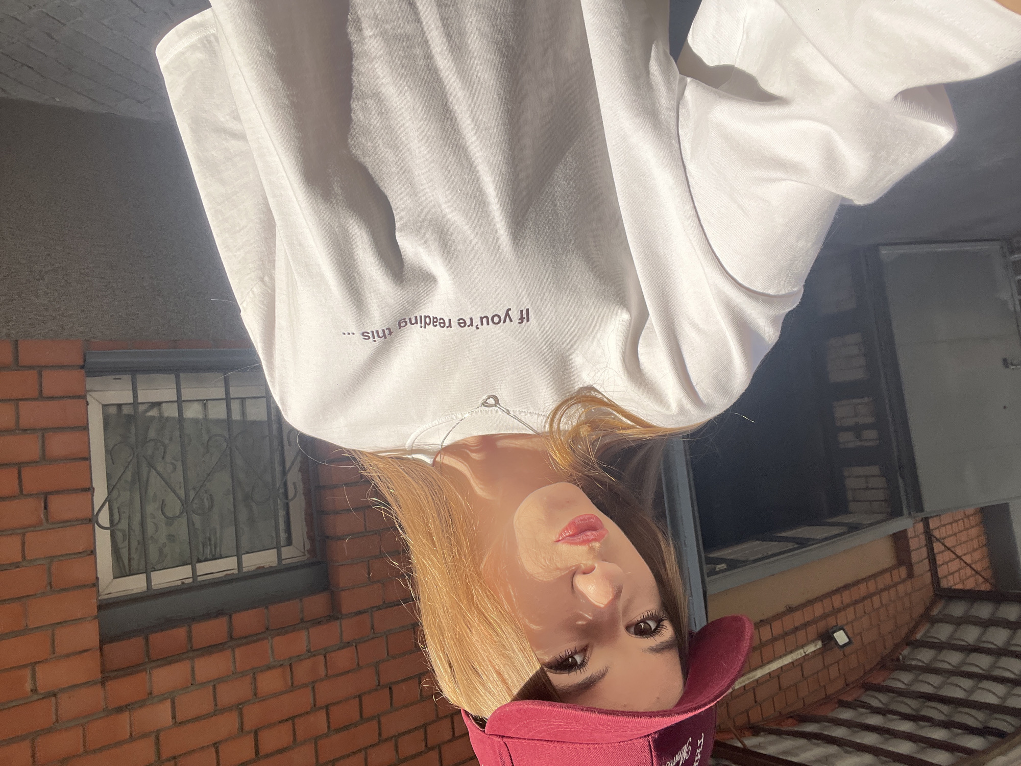 A young woman with blonde hair and red lipstick poses for a selfie outdoors. She is wearing a white t-shirt with the text "If you're reading this..." printed in purple on it. She also wears a dark red baseball cap. The image appears to have been taken during the daytime, with sunlight casting shadows on her face and the white t-shirt.
The background consists of brick walls and a window with a metal grate. The overall setting suggests an urban or suburban environment. The woman's expression is neutral, and the lighting implies it might be a sunny day. The orientation of the photo is unusual, with the subject appearing upside down. The provided location context of Dnipro, Ukraine, places this scene within that city.