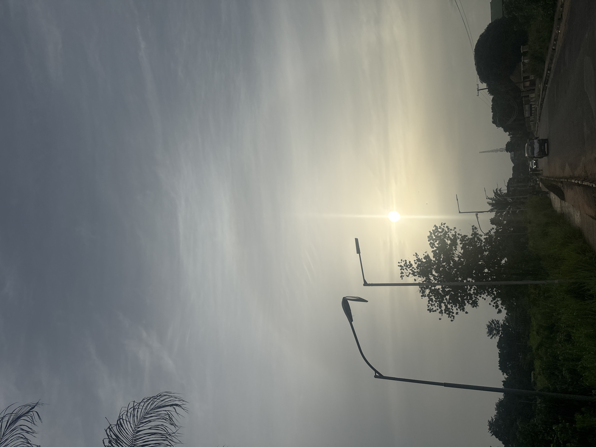 The image captures a street scene in Awka, Nigeria, likely during the late afternoon or early morning, given the diffused sunlight. The sky is overcast with a soft, pale glow emanating from the sun, which appears as a bright, star-like orb low on the horizon, casting a gentle haze over the scene.
Two streetlights are prominently featured in the foreground. The one on the left is taller and has a curved arm, while the one on the right is shorter and more angular. Both are silhouetted against the bright sky. Beyond them, a road stretches into the distance, curving slightly to the right. A few vehicles are visible on the road, including a white minivan in the center and a dark-colored car further down. The road has a paved surface with a white line marking the edge.
To the left of the road, there is a narrow, paved walkway bordered by tall, green grass and vegetation. Several trees are scattered throughout the landscape, with lush green foliage. In the background, more trees and some buildings can be seen, partially obscured by the haze. A tall transmission tower is visible in the far distance, to the right of the road.
On the right side of the road, there is a low concrete wall or curb, with more vegetation and trees behind it. The overall impression is of a semi-urban environment with ample greenery. The lighting suggests a time of day with soft shadows and a muted color palette.
