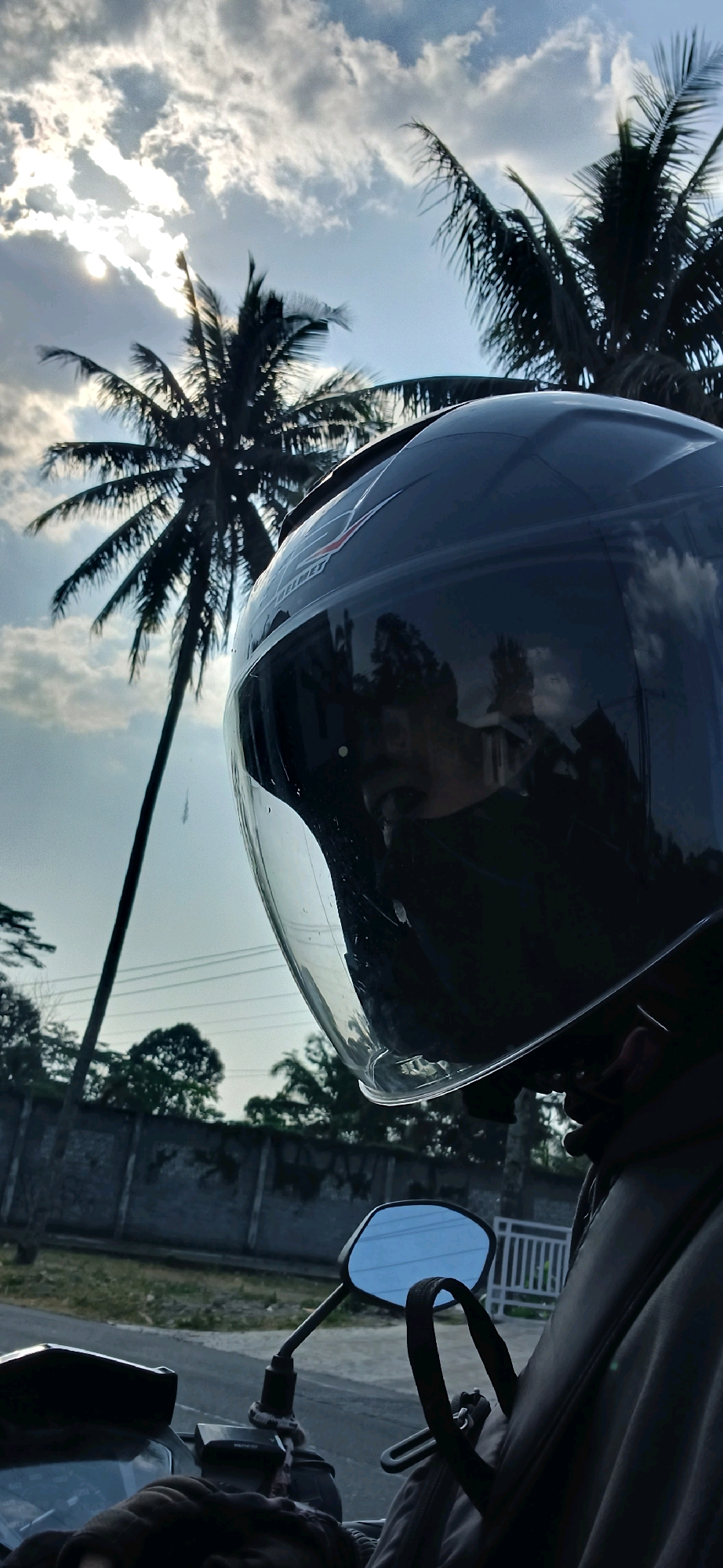 A vertical, eye-level medium shot captures a motorcycle helmet in the foreground, reflecting the sky and a person's face. The helmet is dark and glossy, with a clear visor that is slightly open. Through the visor, the reflection shows a person's eye and part of their face. The background features two tall palm trees silhouetted against a cloudy sky with the sun breaking through. A low stone wall and some greenery are visible below the trees, suggesting an outdoor setting. The image appears to be taken in Tengahan, Indonesia, as indicated by the presence of palm trees and the overall tropical atmosphere.