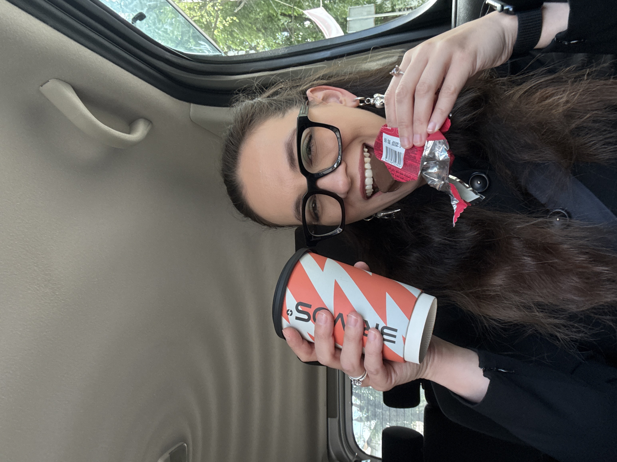 A woman with long, dark brown hair and black-rimmed glasses is sitting in the passenger seat of a car, holding a coffee cup and a chocolate bar. She is smiling and taking a bite out of the chocolate bar. The coffee cup is orange and white with a black lid. She is wearing a black jacket and jewelry on her hands and ears. The interior of the car is visible, including the ceiling, a handle, and a window. Outside the window, trees and a flag are visible, suggesting a suburban or park-like setting. The image appears to be taken in Chaikovsky, Russia, based on external context.