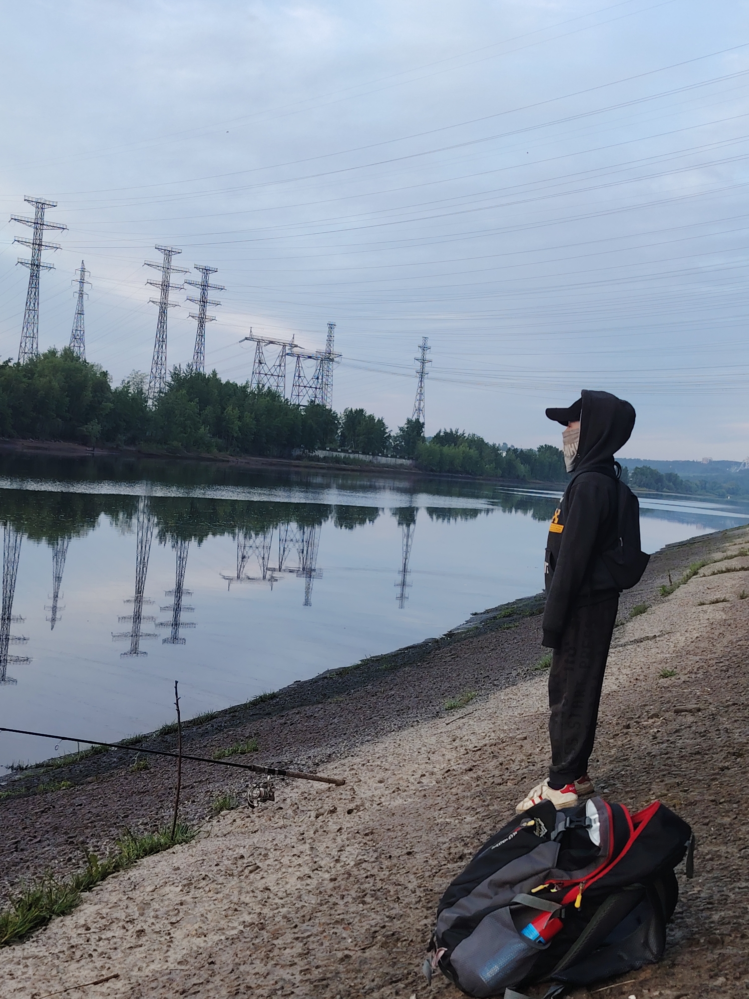 This image depicts a single individual standing on a concrete riverbank, gazing out across a wide, calm body of water. The person is dressed in a black hooded sweatshirt, dark pants, and a baseball cap, and is wearing a light-colored face mask. A multi-colored backpack (black, grey, and red) is placed on the concrete next to their feet. To the left, a fishing rod is set up, supported by a stick, with its line extending into the water, indicating fishing as the primary activity or intention.
The scene is characterized by a tranquil river or canal, whose still surface beautifully reflects the overcast sky and the structures on the opposite bank. The sky is a uniform, light grey, suggesting either early morning or late evening light. On the far bank, a line of green trees runs along the water's edge, punctuated by several large, metallic power line towers (electricity pylons) that stand tall against the skyline, their reflections clearly visible in the water below. These power lines and towers are a prominent feature, creating an industrial backdrop to the natural riverine environment. The setting is in Chaikovsky, Russia. No discernible text is clearly visible on clothing or objects within the frame.