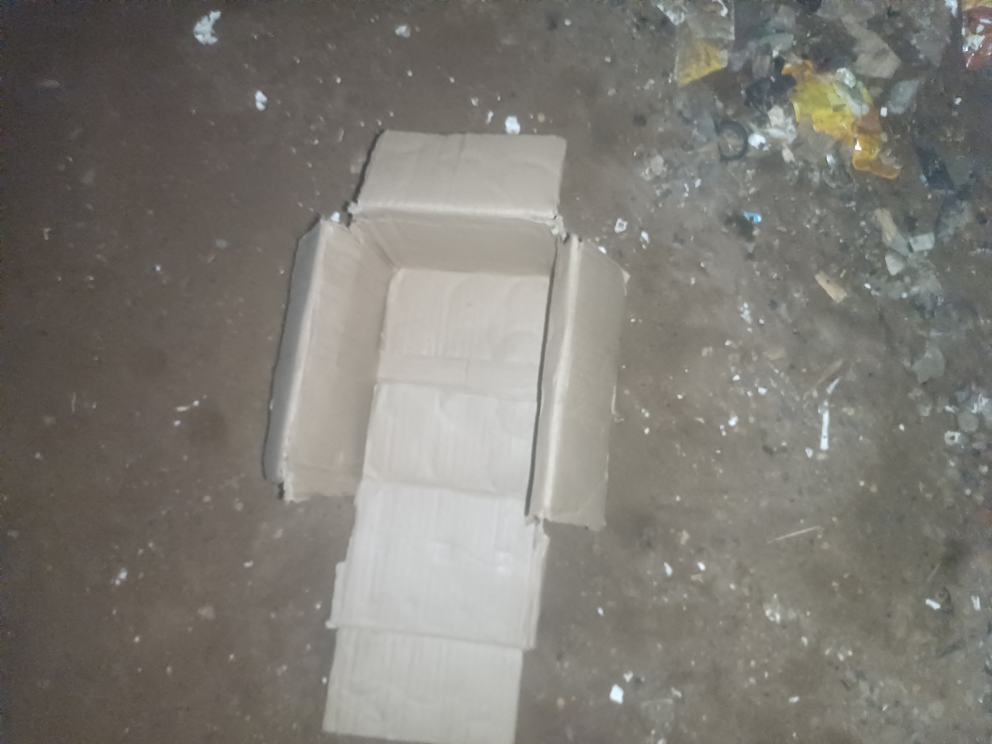 A cardboard box is partially unfolded and lying on the ground. The box is beige and appears to be made of corrugated cardboard. It is open on the top, with its sides folded down. The ground beneath the box is a mixture of dirt and debris, including small white specks, some larger greyish fragments, and scattered pieces of what appears to be trash, including yellow and orange plastic. The overall scene suggests an outdoor environment, possibly an alleyway or a neglected area within Wukari, Nigeria. There are no people visible in the image.