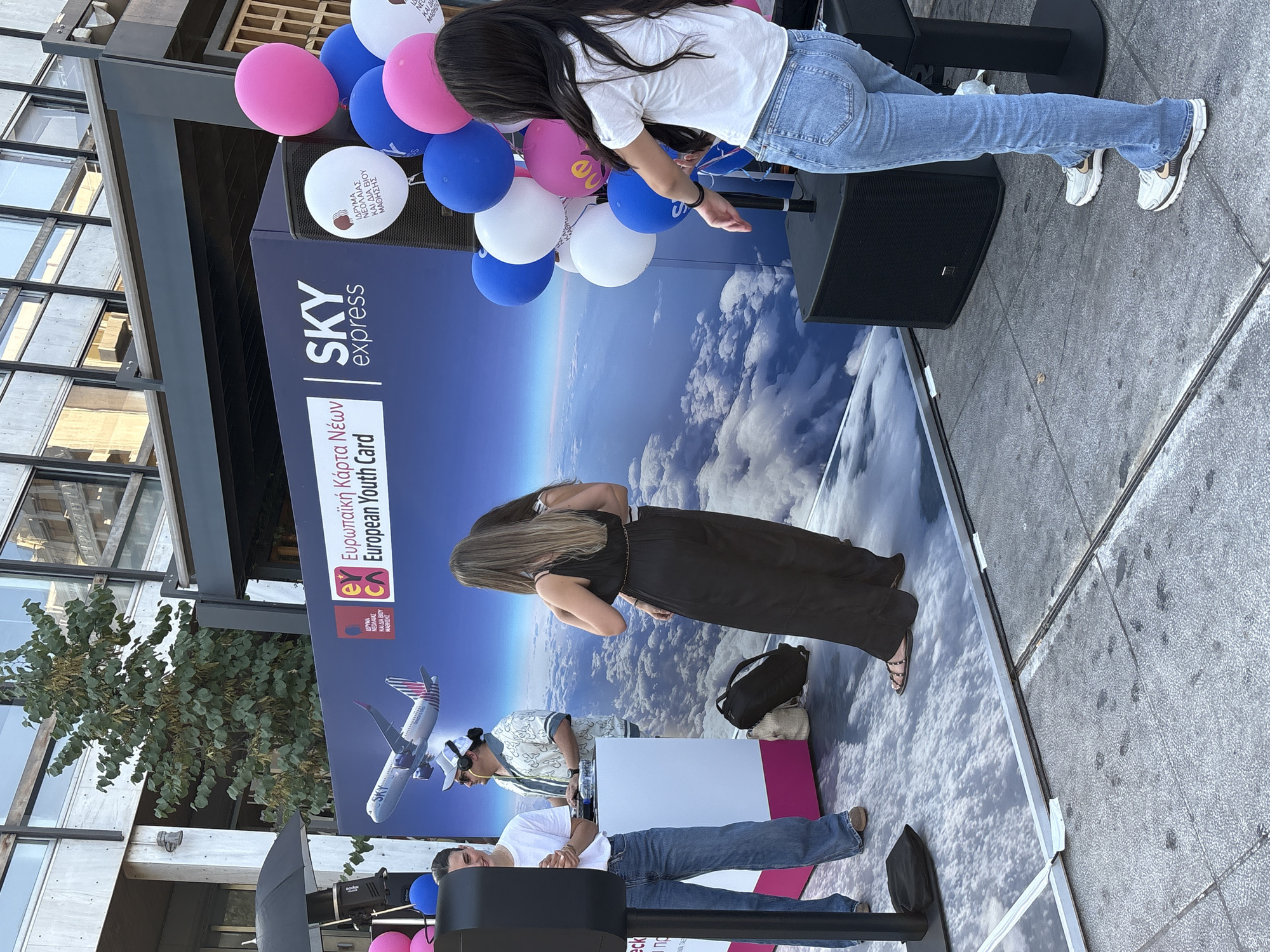 In this image, several people are gathered outdoors, seemingly at an event. The background features a large banner with the "SKY express" logo and the text "European Youth Card" in both Greek and English, suggesting a promotional or informational event related to youth travel or opportunities. Balloons, predominantly pink and blue, are present, adding a festive atmosphere.
One woman, wearing a white t-shirt and blue jeans, is bending over, possibly interacting with something near a speaker or stage. Another woman in a black dress is also seen from behind, with long brown hair. In the foreground, two men are visible. One, wearing a white t-shirt and blue jeans, is seated or leaning. Another man, wearing a light-colored patterned shirt and a baseball cap, stands behind a white counter.
The setting appears to be a public space, possibly a plaza or street, with buildings and trees visible in the periphery. The ground is paved with grey tiles, and the lighting suggests it is daytime, likely sunny. The overall mood seems lively and engaging, typical of a public event. The presence of promotional materials and the gathering of people indicate an ongoing activity. The text on the banner is clearly legible, providing context about the event's purpose.