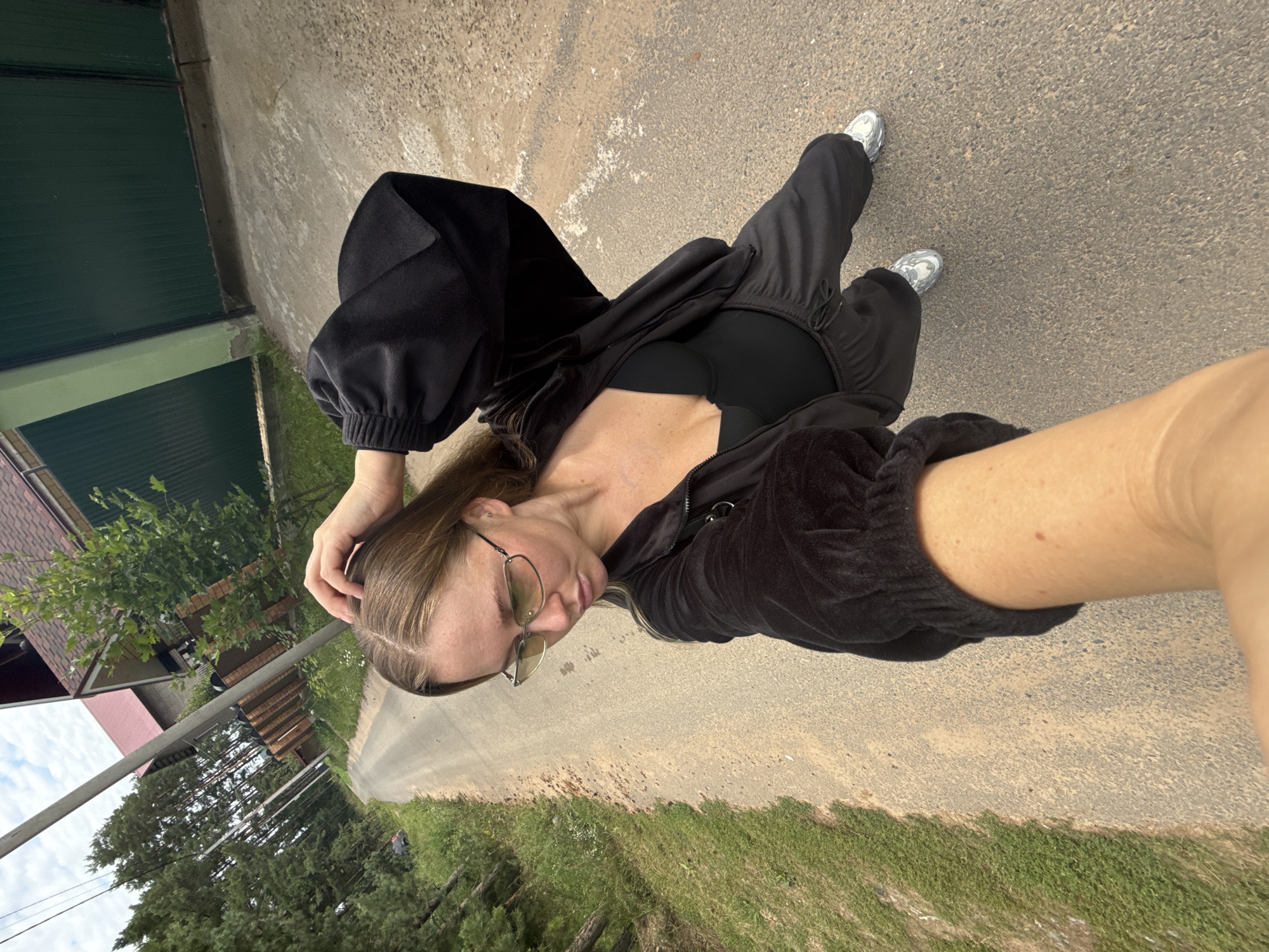 A young woman with long, light brown hair and glasses is standing on a rural road in Chaikovsky, Russia. She is wearing a black velour tracksuit, a black bodysuit, and silver sneakers. The woman is holding her left hand to her head and looking down. The road is paved with asphalt and is bordered by trees and grass on either side. In the background, there are houses and a fence. The sky is cloudy.