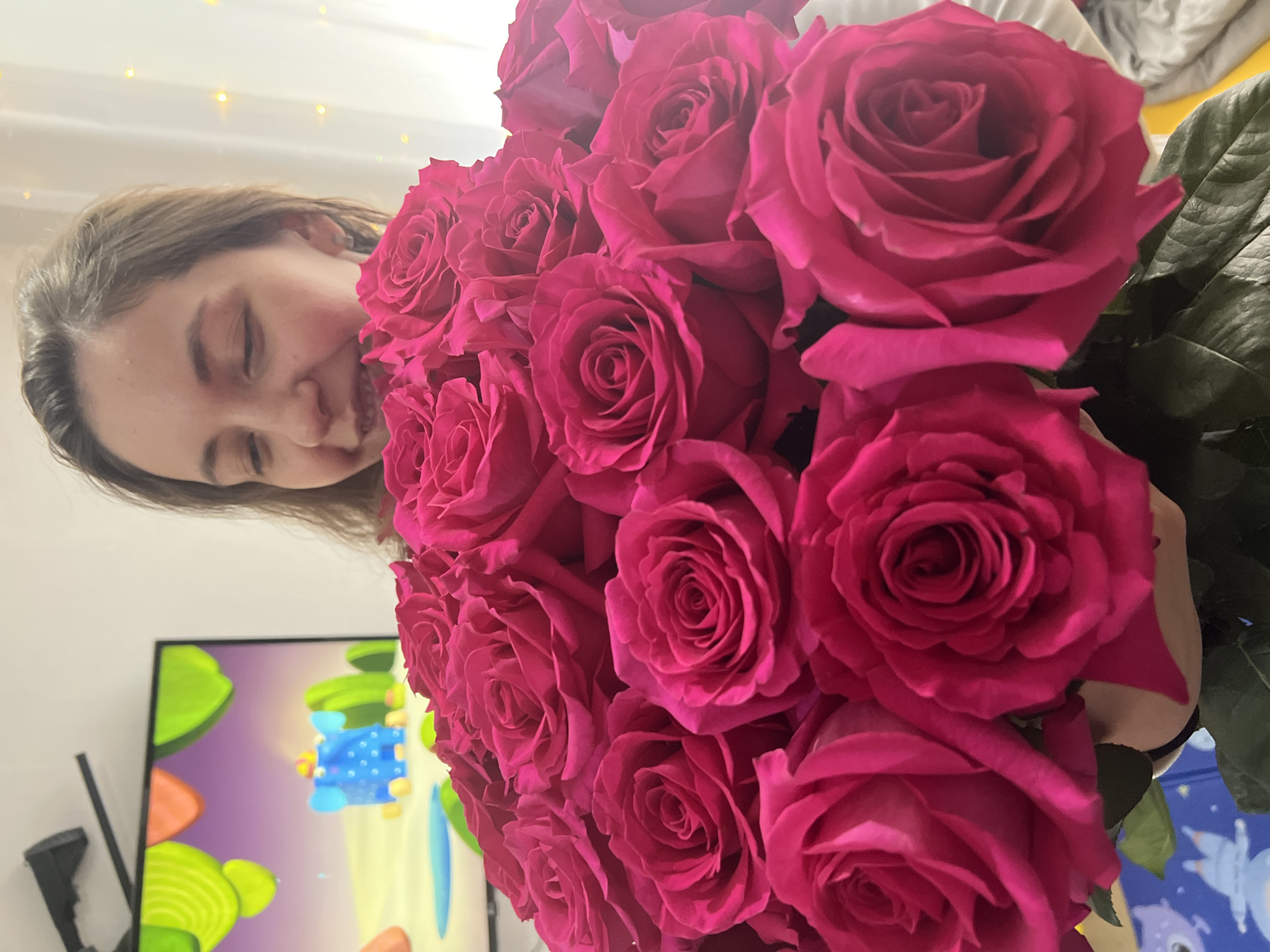 A young woman holds a large bouquet of vibrant pink roses, her face close to the flowers and eyes closed as if enjoying their scent. The background is a brightly lit room. A television screen displaying a colorful cartoon is visible in the lower-left corner. The location is not possible to determine based on the provided image.