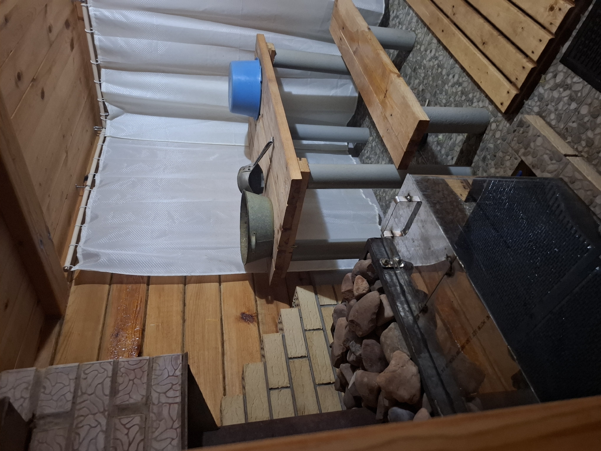 The image shows the interior of what appears to be a sauna or banya. The walls are lined with light-colored wood paneling, and a white curtain is draped along one wall. A wooden structure with gray pipes and wooden planks serves as a seating arrangement or part of the sauna's infrastructure. A blue bucket and a metal dipper are attached to the structure. To the right, there's a stone-filled container, suggesting a traditional sauna stove. The floor is a mix of wooden planks and tiled surfaces. The overall setting evokes a warm, rustic atmosphere. The location is Амзя, Russia.