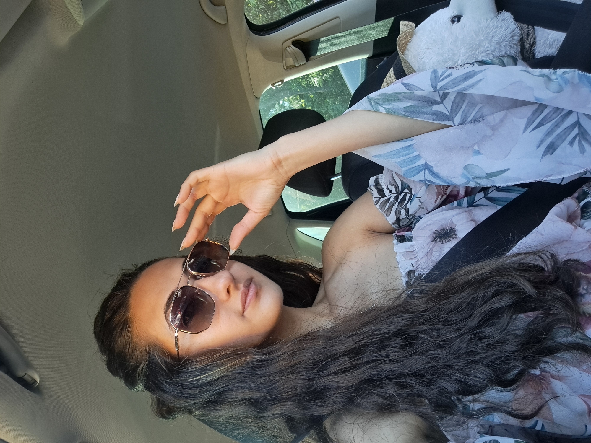 A young woman with long, dark, wavy hair is sitting in the passenger seat of a car. She is wearing large, aviator-style sunglasses with gold frames and an off-the-shoulder top with a floral print. Her right hand is holding the sunglasses up to adjust them. A seatbelt is visible across her chest. Beside her, a white stuffed animal wearing a straw hat is secured with a seatbelt. The interior of the car is visible, including the headrest, ceiling, and a portion of the window showing greenery outside. The image appears to be taken from a slightly elevated angle, looking down at the woman.