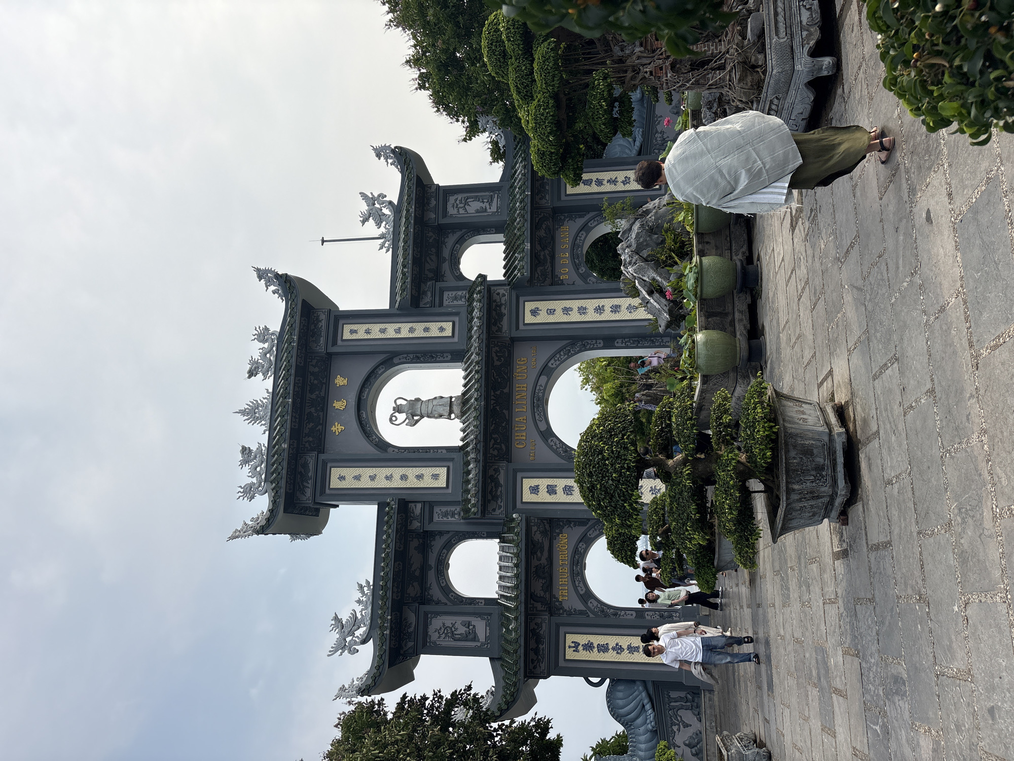 This image captures the entrance to the Linh Ứng Pagoda, located in the city of Đà Nẵng, Vietnam. The pagoda is fronted by a large, ornate, multi-tiered stone gate adorned with traditional Vietnamese architectural elements, including dragon and phoenix motifs on its roof edges. The gate features several arched openings and is inscribed with gilded Vietnamese characters. A statue is visible within one of the upper archways. Lush bonsai trees and potted plants flank the gate.
Several people are visible. A person in a light-colored shirt and green skirt is standing near the front, possibly admiring the architecture. Further back, other individuals are walking through the entrance, suggesting that the pagoda is open to visitors. The ground is paved with stone tiles. The sky is overcast.