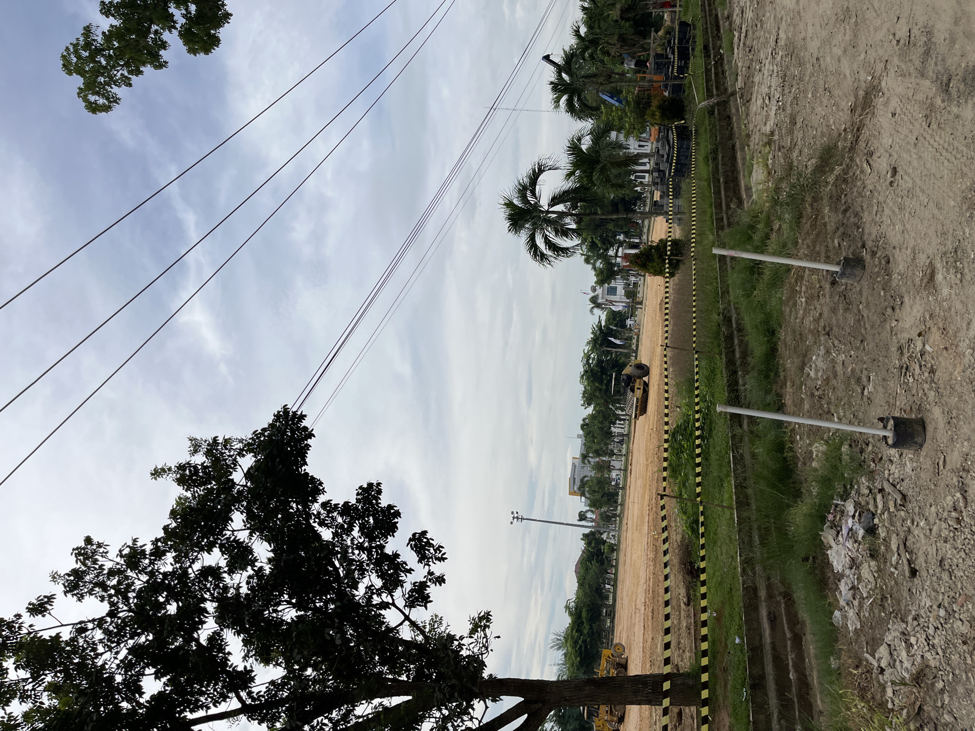 This media file shows an outdoor scene at a construction site in Bundar, Indonesia. The image is oriented vertically, with the sky occupying the upper portion and the ground the lower. Several overhead power lines stretch across the frame.
In the foreground, a silhouetted tree with dense foliage is on the left. The ground appears to be a mix of dirt, gravel, and some patches of grass. Yellow and black striped caution tape runs along the edge of a pathway or excavated area. Several utility poles with lights are visible, one of which is in the mid-ground to the right, with a black base and a silver pole. A pile of trash and debris is scattered near this pole.
Further into the background, a large area of cleared and leveled earth is visible, suggesting ongoing construction. There are palm trees and other vegetation interspersed with buildings that appear to be houses or smaller structures, some with white exteriors. Construction equipment, specifically yellow bulldozers or similar machinery, can be seen in the mid-ground and further back. A tall light pole is also present in the distance. The sky is overcast with a mix of white clouds and a pale blue hue, indicating it is likely daytime, possibly morning or afternoon, with cloudy weather. There is no visible text in the image. The overall impression is one of a developing area, possibly a housing or commercial project, with elements of infrastructure and construction activity.