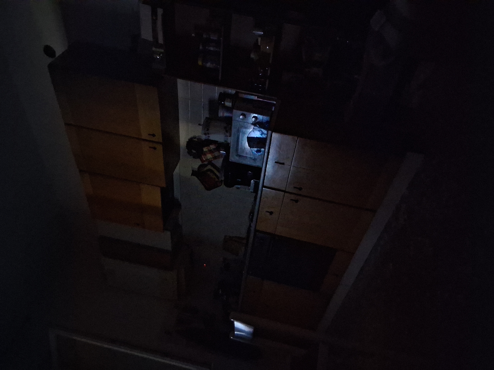 The image depicts a dimly lit kitchen, likely in an apartment in Colorno, Italy. The scene is largely obscured by shadow, but visible elements include wooden kitchen cabinets, a countertop, and various kitchen appliances and objects. In the center of the frame, a microwave oven is visible on the countertop, with a light emanating from within. To the right, a shelf unit holds several jars and containers. The overall impression is of a functional but somewhat cluttered kitchen, appearing undisturbed. No individuals are present in the image.