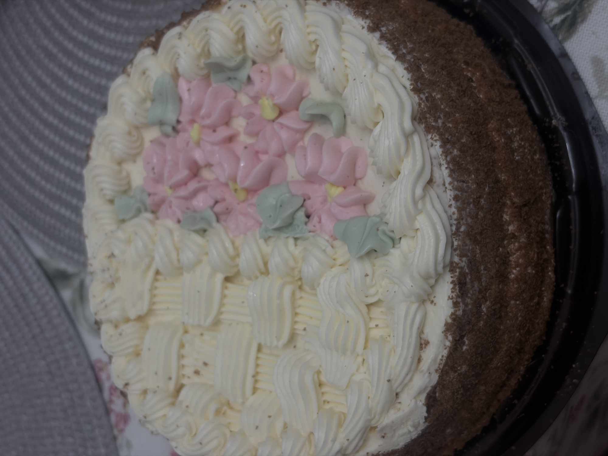 Here is a detailed description of the image:
The image shows a close-up view of a decorated cake. The cake is round and appears to be sitting in a black plastic container. The sides of the cake are covered in dark brown crumbs, likely chocolate. The top of the cake is decorated with white buttercream frosting. The frosting is piped in various patterns, including swirls and basketweave designs. In the center of the cake, there are pink buttercream flowers with light green leaves and tiny yellow stamen. The frosting details appear handcrafted. The background of the image shows a slightly blurred gray and white patterned cloth. There are no people in the image. The location is unknown.