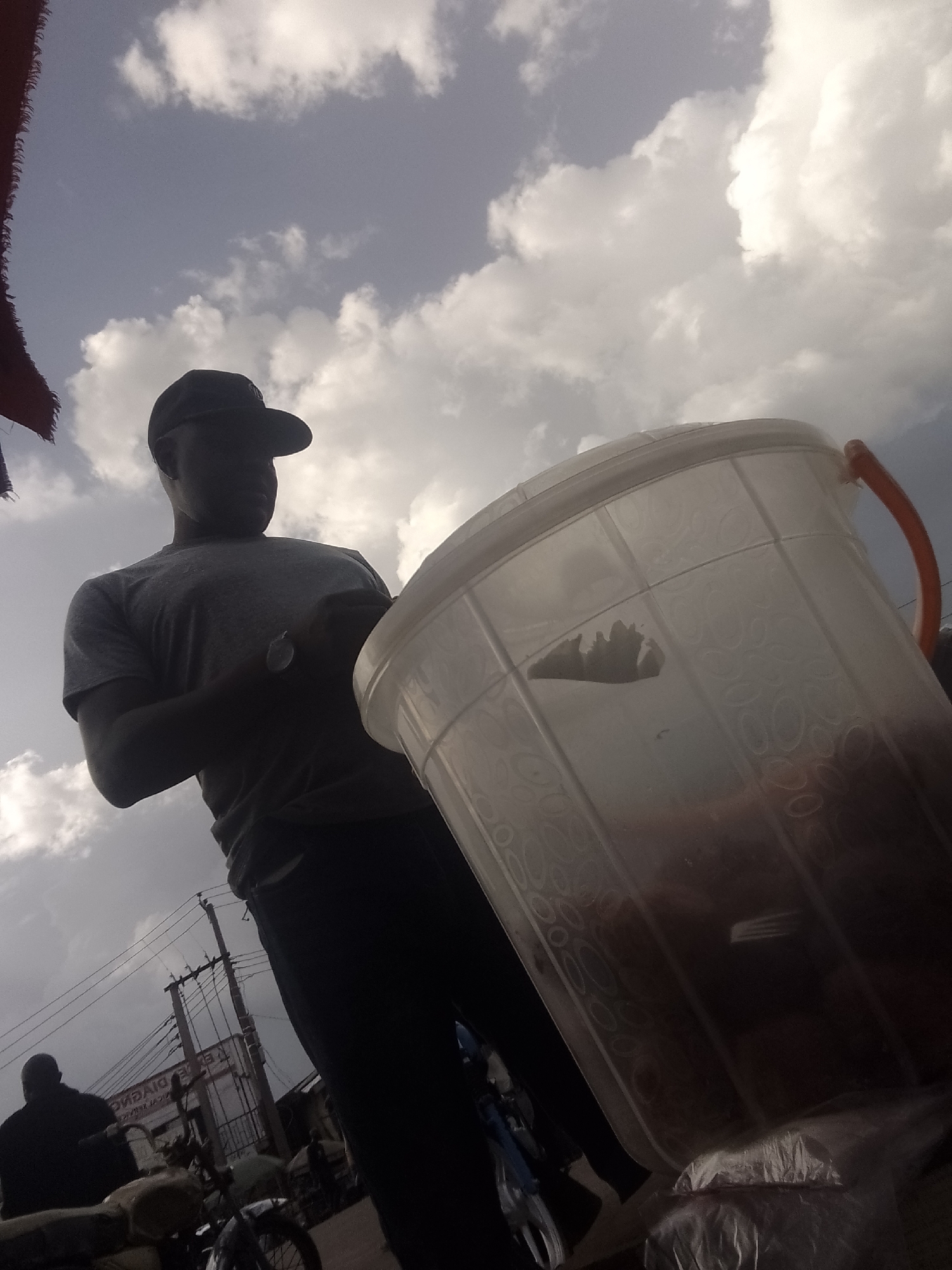 The image shows a silhouette of a person wearing a baseball cap and a t-shirt, standing next to a large, transparent plastic container with an orange handle. The container appears to be filled with a dark liquid or contents. The person is looking down and appears to be interacting with the container, possibly serving from it.
The setting is outdoors, indicated by the sky and clouds visible in the background, suggesting it is daytime, possibly late afternoon or early evening given the lighting. The sky is partly cloudy with bright white clouds against a bluish-grey sky. There are some electrical poles and wires in the background, along with what appears to be a building or storefront with some faded text that is difficult to read. In the lower part of the image, there are glimpses of a motorcycle and another person in silhouette.
The overall impression is of a casual outdoor scene, possibly a market or a street vendor's stall. The low angle of the shot and the silhouette of the main subject create a somewhat dramatic or moody atmosphere. There is no visible text that can be clearly deciphered.